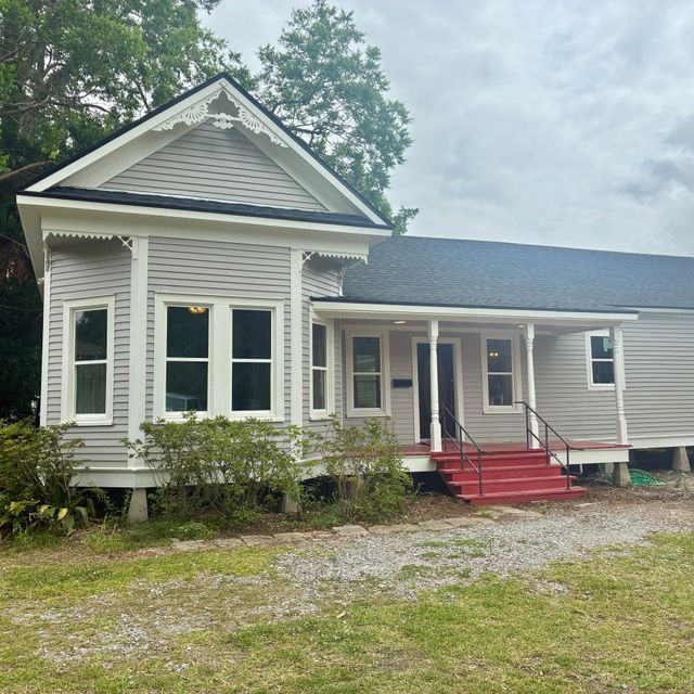 White Victorian-style house with red front steps, covered porch, and gravel driveway under cloudy sky