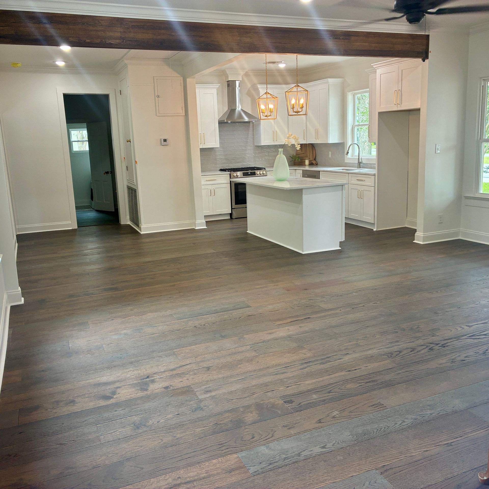 Spacious empty living room with dark wood floors, white walls, and a bright kitchen with island and chandelier.