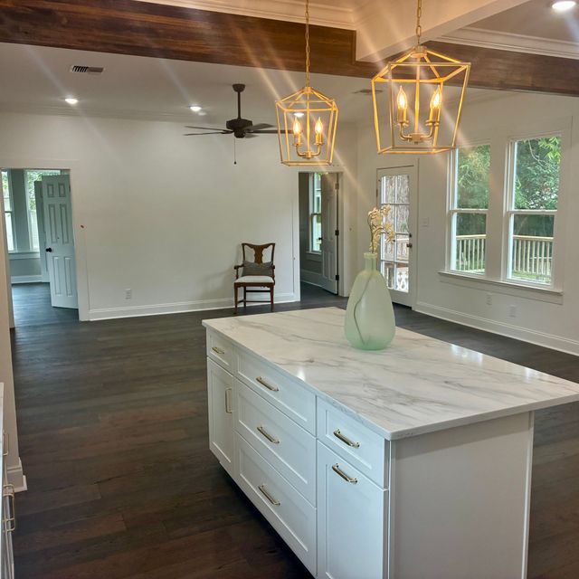 Bright kitchen with marble island, brass pendant lights, dark floors, and a view into an adjacent room through windows.