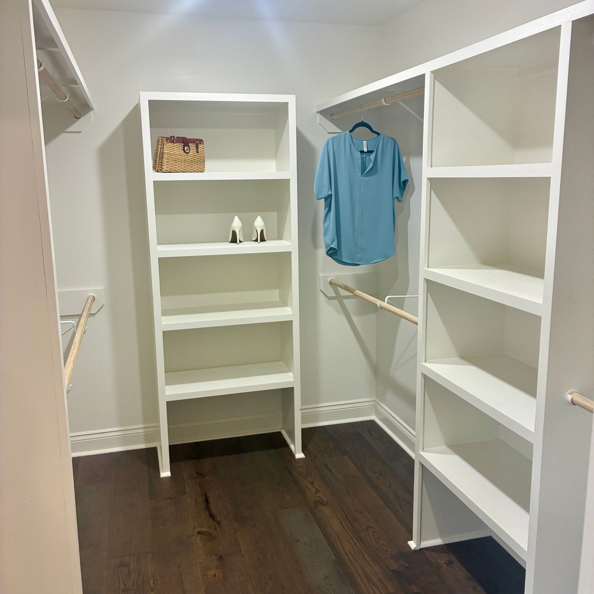 Walk-in closet with white shelves, hanging rods, and dark wood floor; blue shirt on right side
