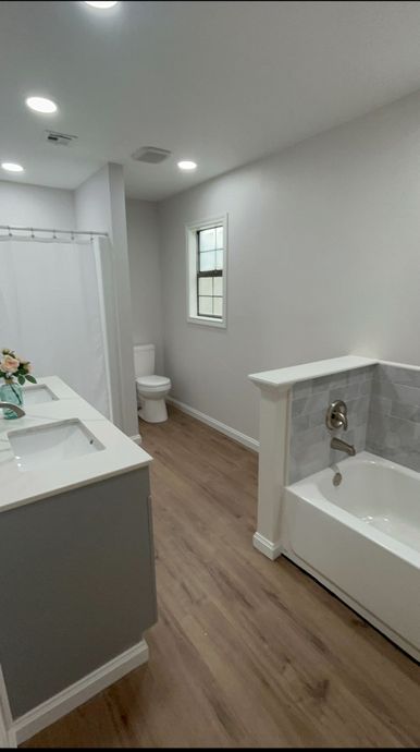 Bright white bathroom with wood floor, vanity, toilet, window, and bathtub/shower area.