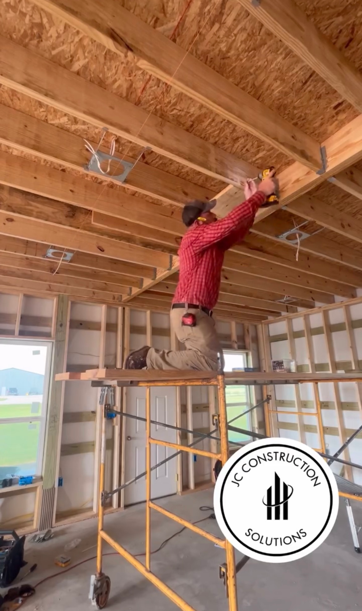 Worker in red hoodie kneels on scaffolding, installing insulation in a framed house ceiling.