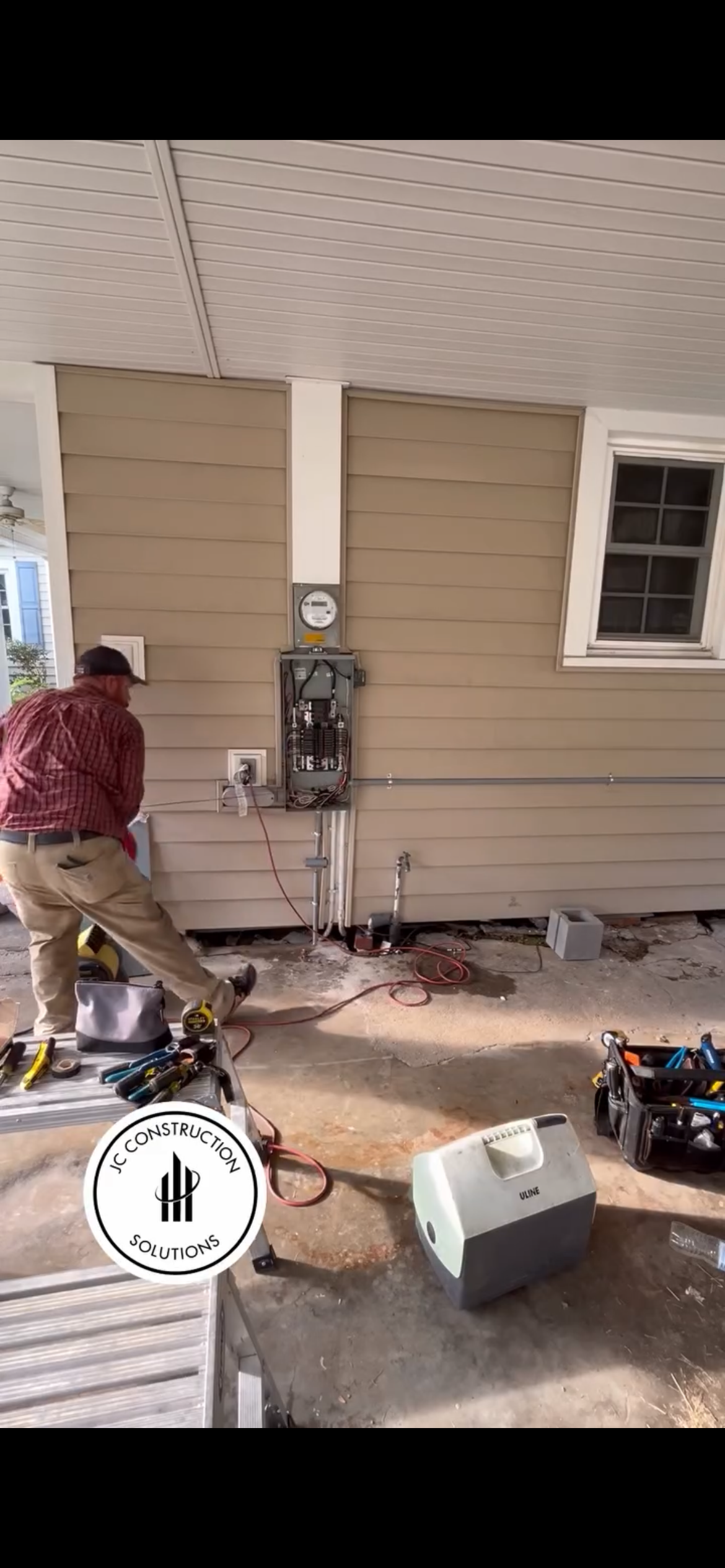 Person working with tools on an exterior house wall beside exposed wiring and equipment.