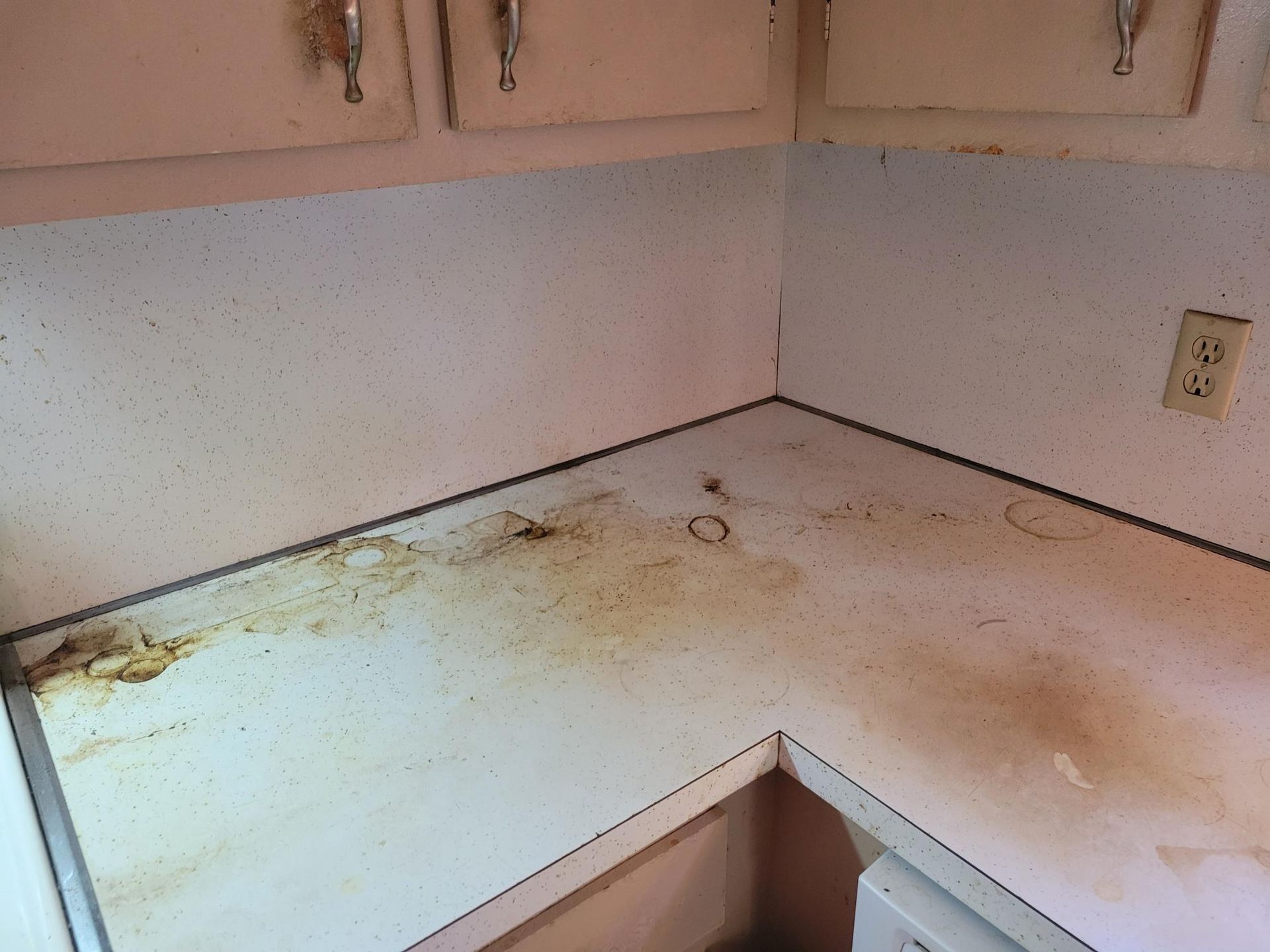 Dirty white kitchen countertop with brown stains, next to white cabinets and a wall outlet.