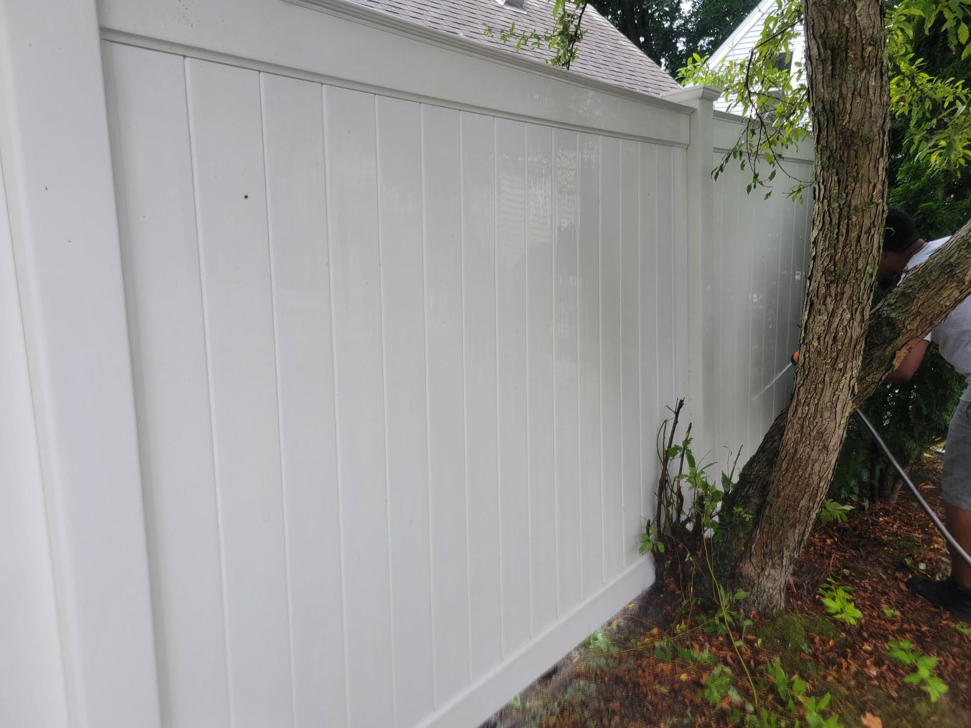 White vinyl fence being power washed by a person near a tree, outdoors.