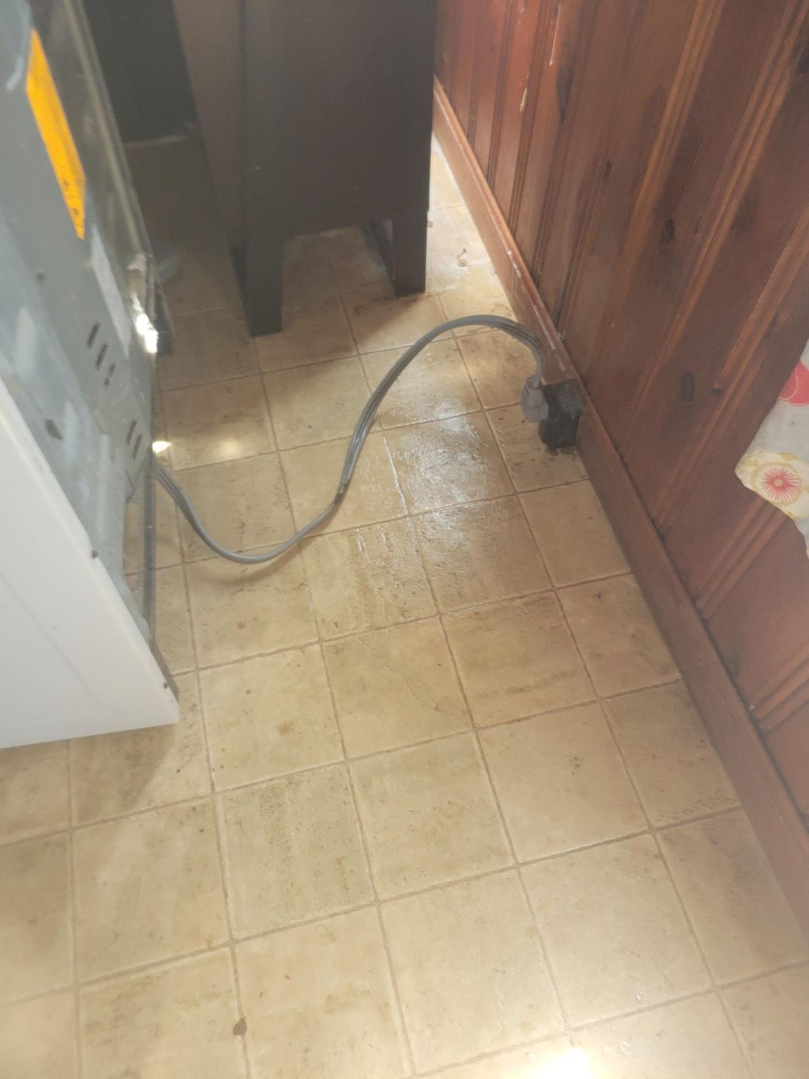 Water on a tiled floor near a power cord and a cabinet, next to wood paneling.