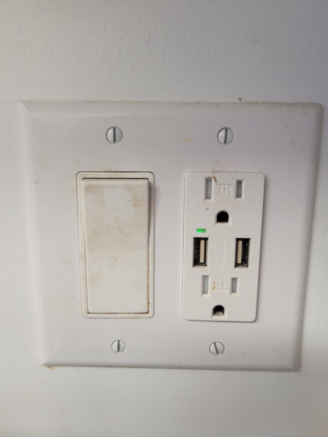White electrical outlet and light switch on a white wall. The outlet includes USB ports, with a green light illuminated.