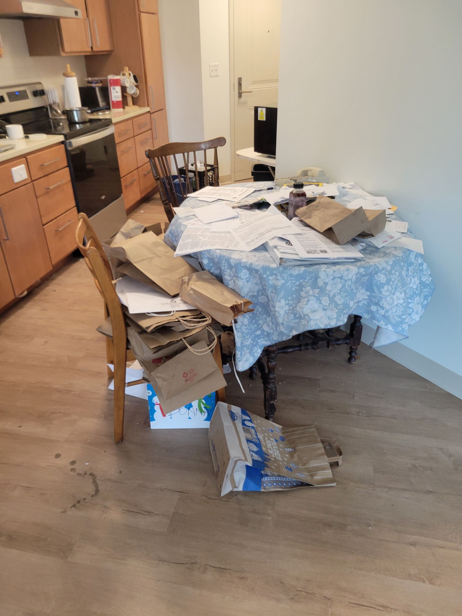 A cluttered kitchen/dining area with a table covered in papers and cardboard. Wooden chairs and cabinets are visible.