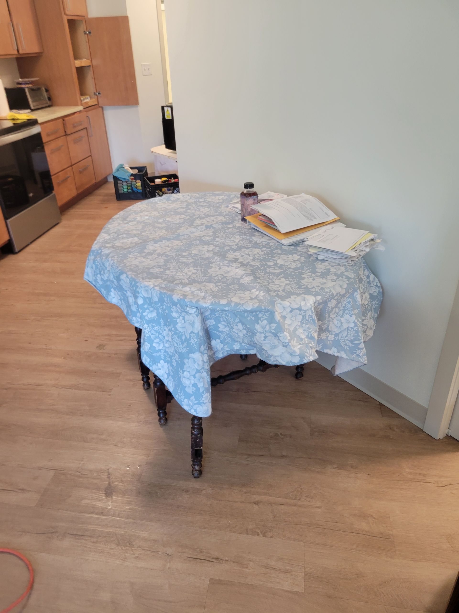 A round table covered with a blue floral tablecloth, with papers and a bottle on top, in a room.