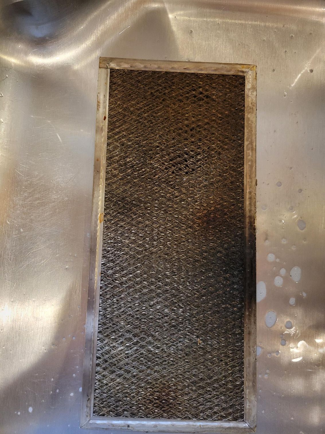 Dirty, rectangular metal range hood filter in a sink, coated in grease.
