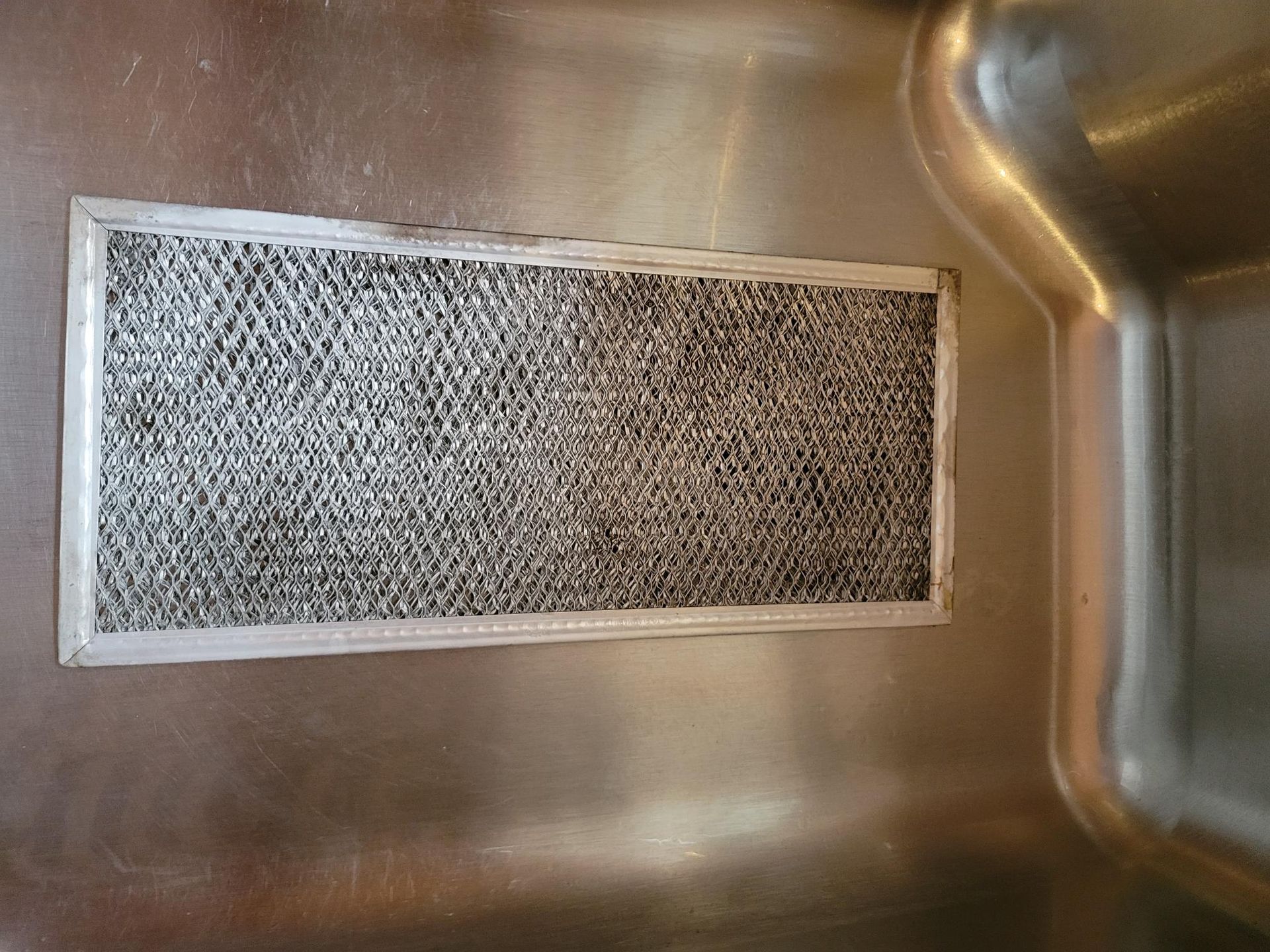 Stainless steel kitchen range hood filter, covered in grease, inside a sink.