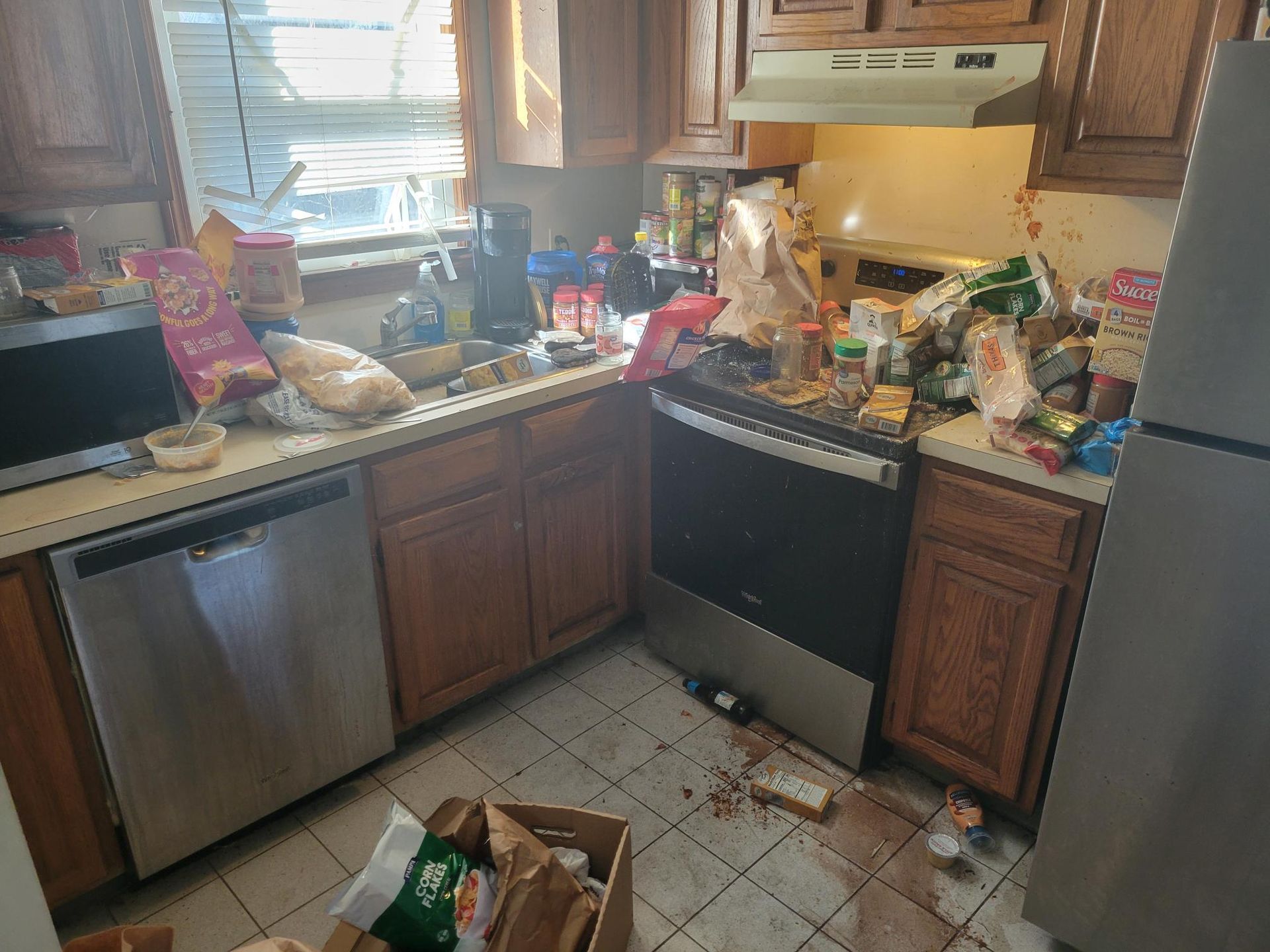 Messy kitchen with counters cluttered with food items, appliances, and spilled liquid on the floor.