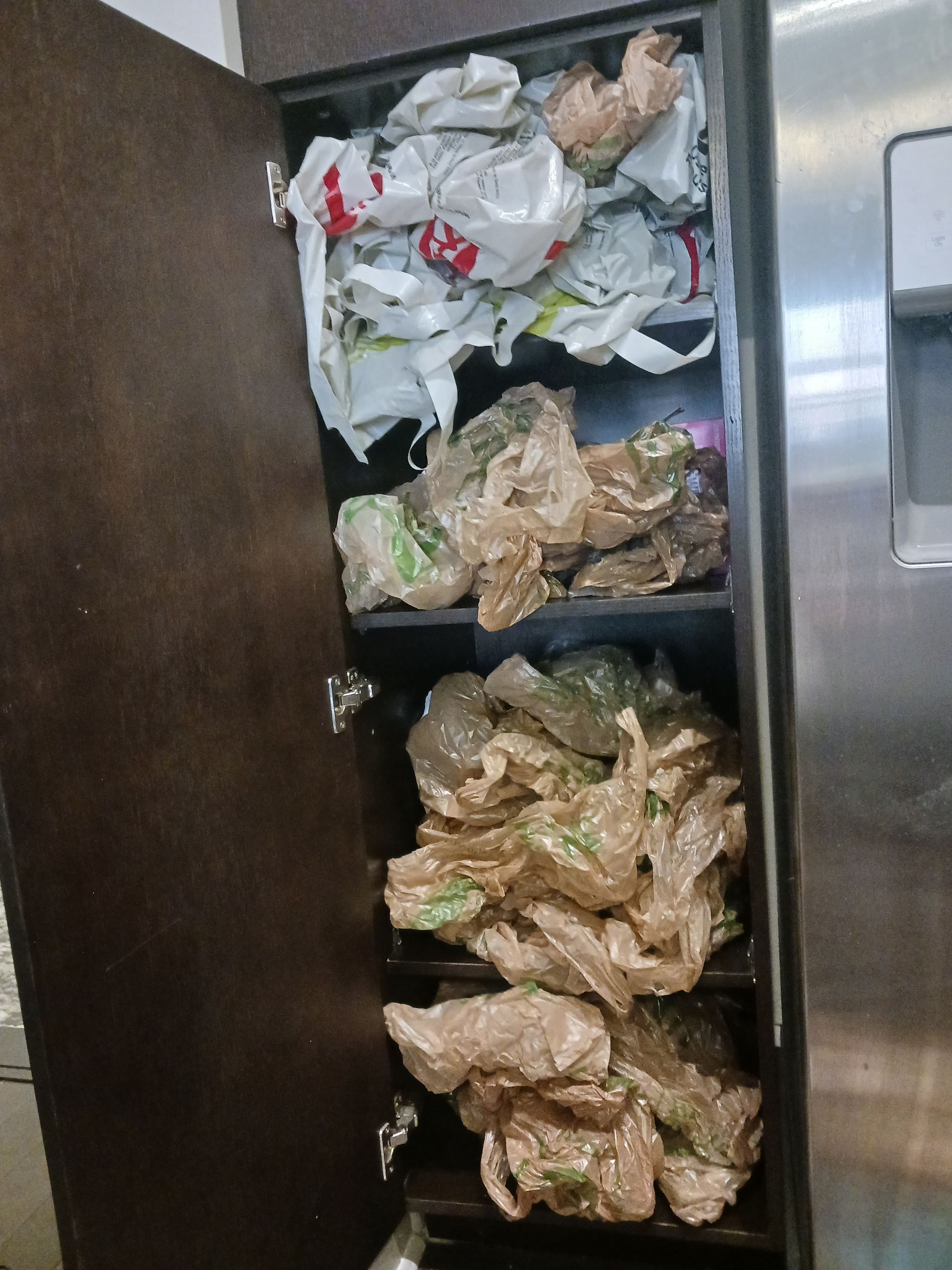 A dark cabinet is overflowing with plastic grocery bags of various colors.
