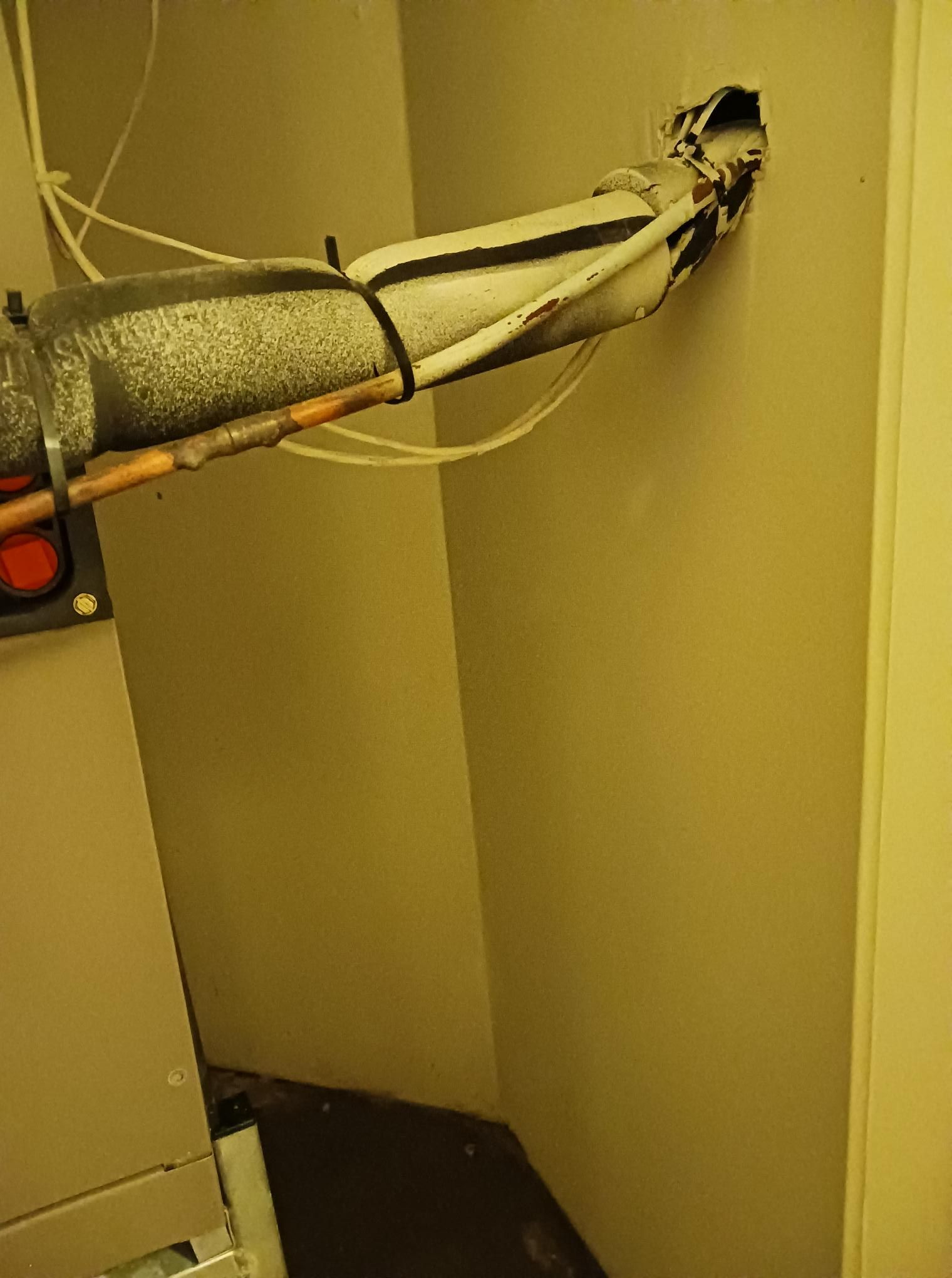 Pipes with insulation emerge from a wall into a corner. Wires run alongside the pipes near a mechanical unit.