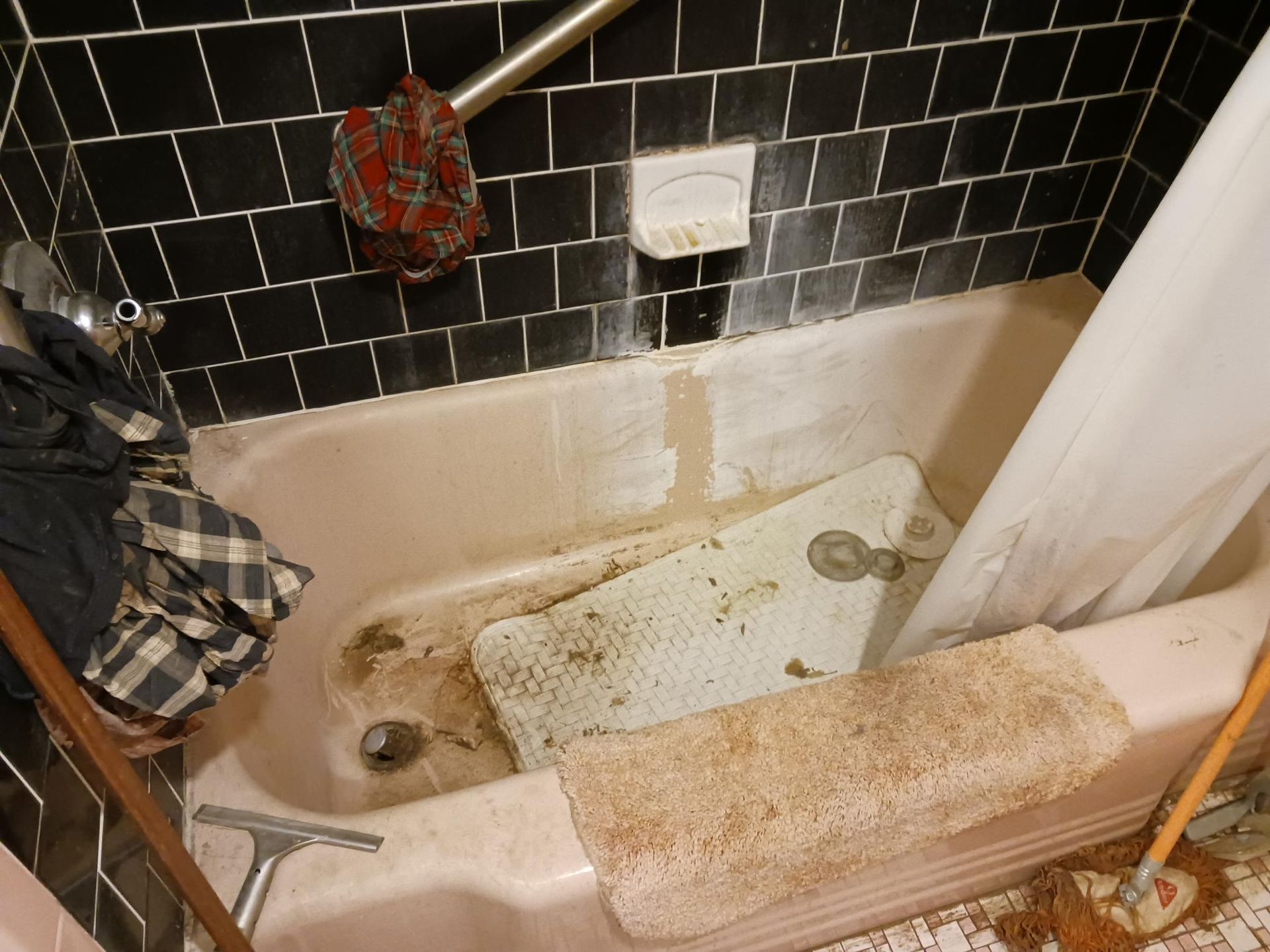 Dirty bathtub with dark tiled walls. Discarded mats, a shower curtain, and various debris are inside.