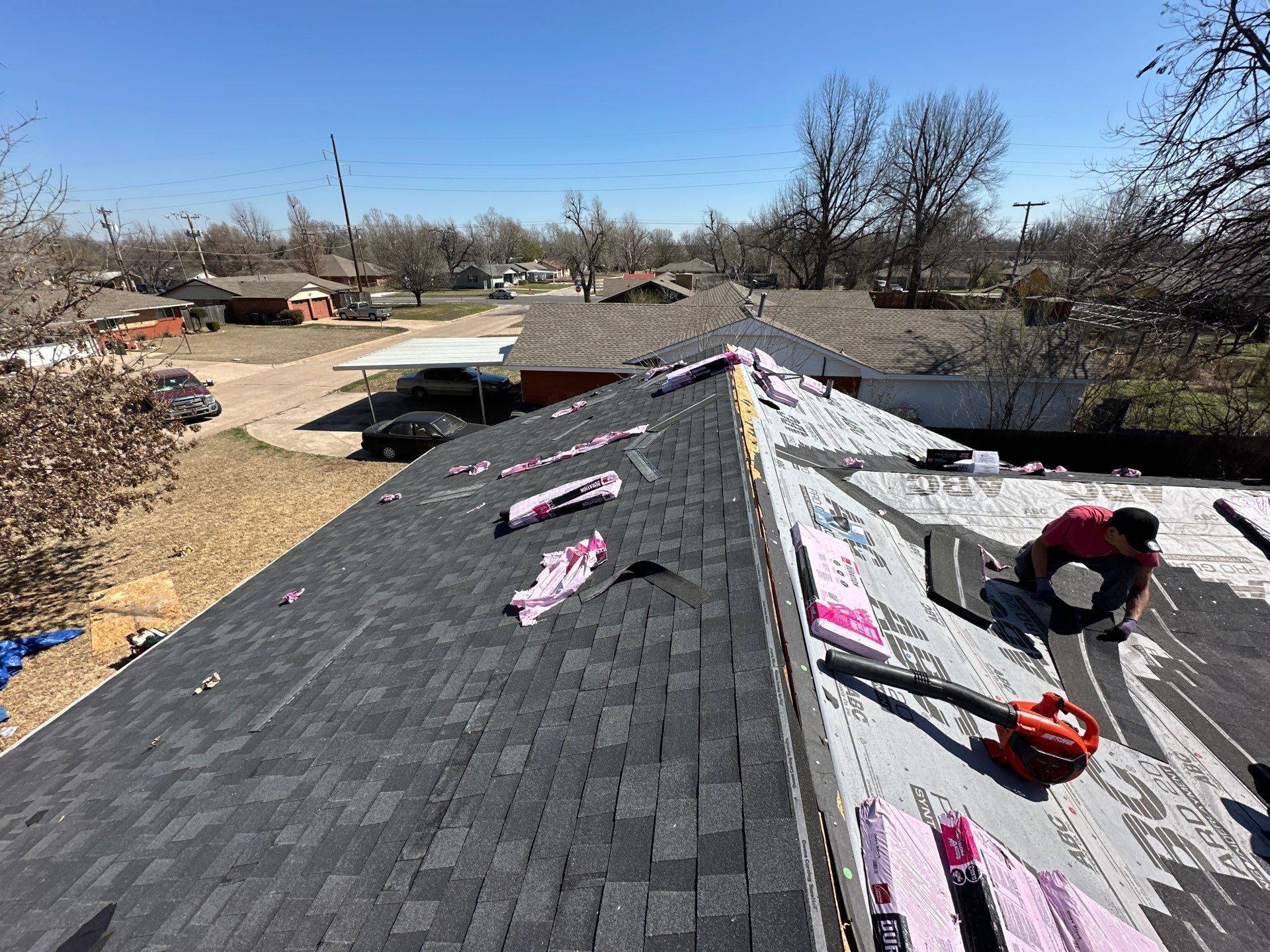 Roofer removing old shingles from a house, with others partially removed, tools present. Blue sky, suburban setting.