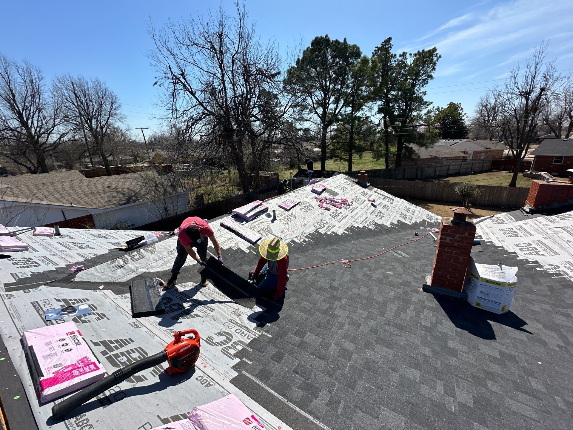 Two roofers working on a roof with asphalt shingles, using tools, surrounded by pink insulation, on a sunny day.