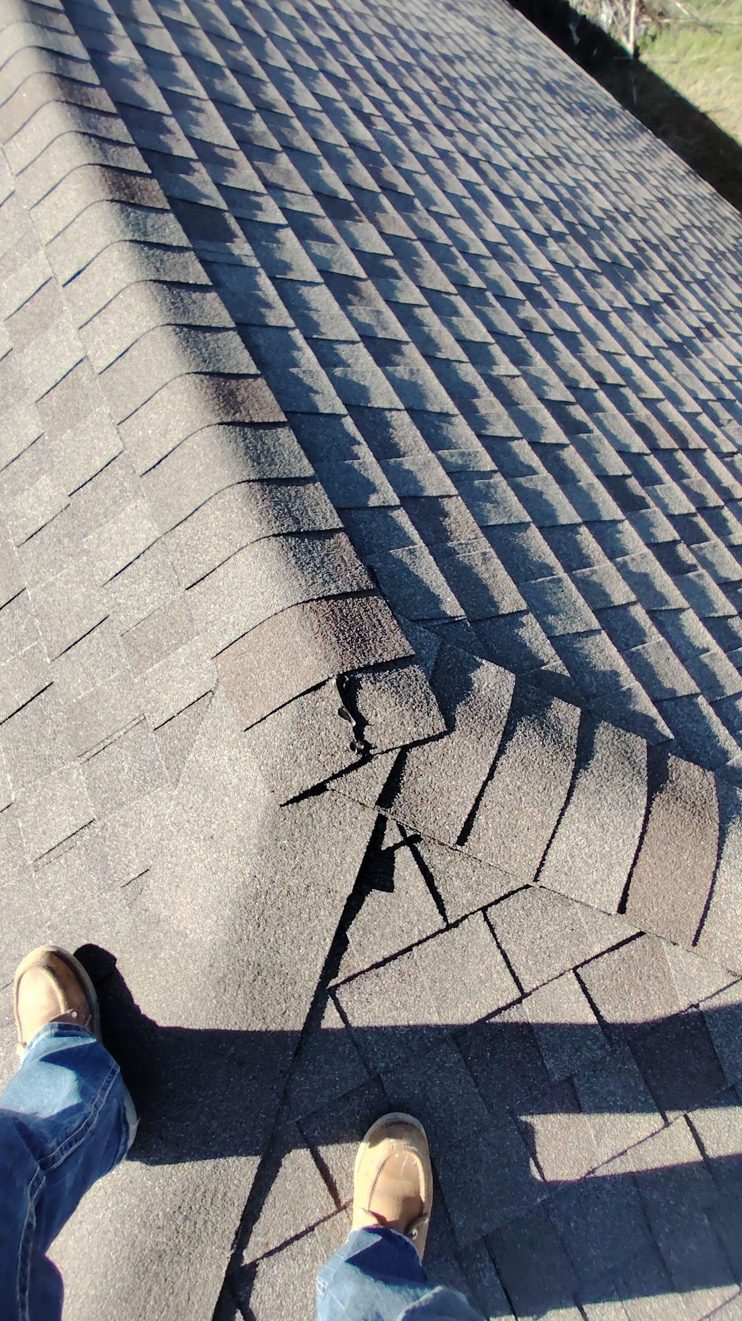 A person standing on a roof with damaged asphalt shingles near a seam. The shingles are grey and textured.