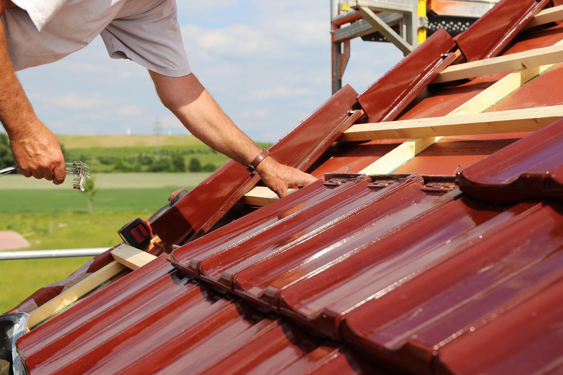 Roofer installing red roof tiles on a sloped roof with wooden supports, sunny outdoor setting.