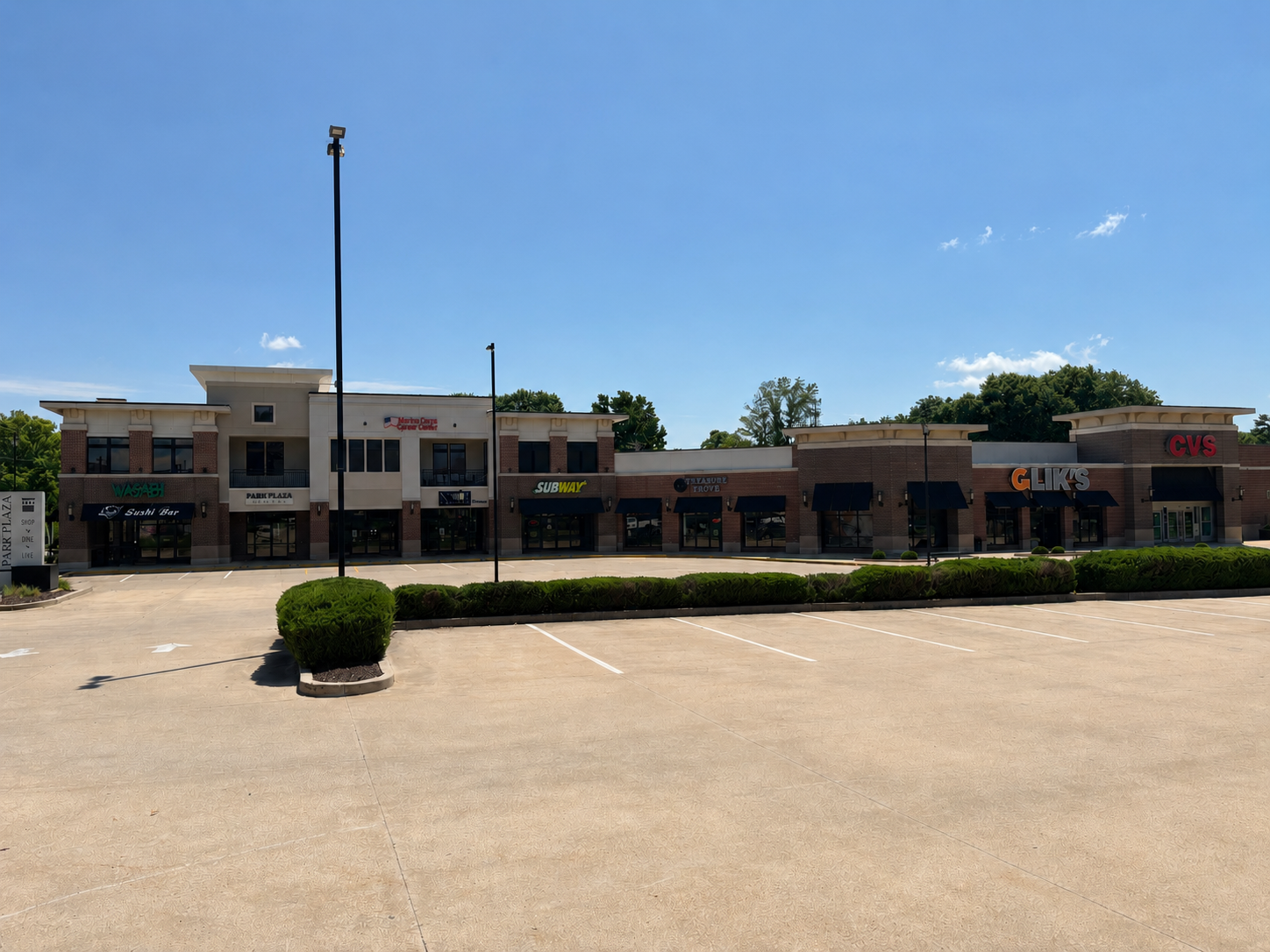 Strip mall storefronts under a bright blue sky, with empty parking lot in front