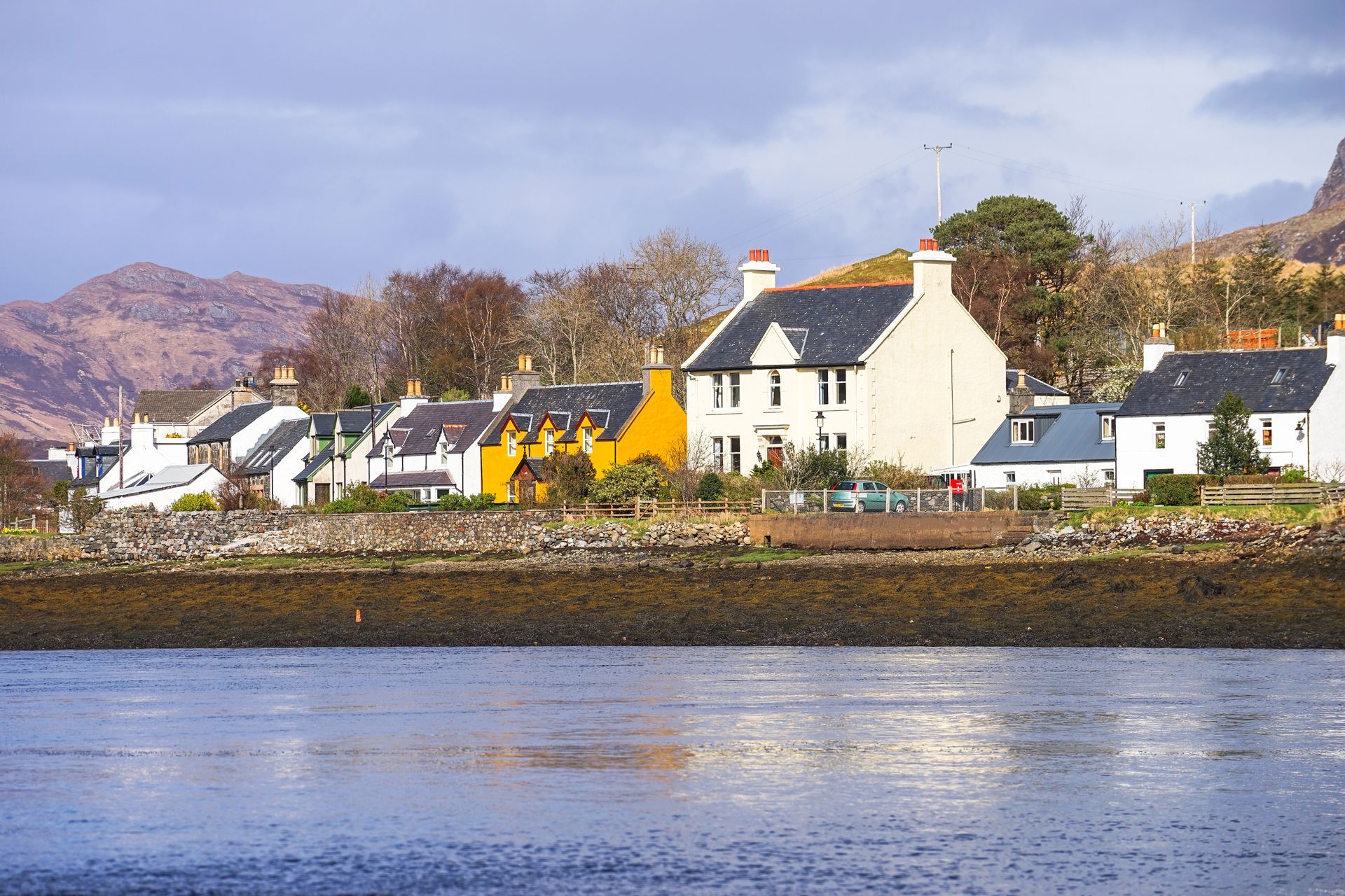 Coastal village with white houses and a yellow building beside calm water, backed by hills and a cloudy sky