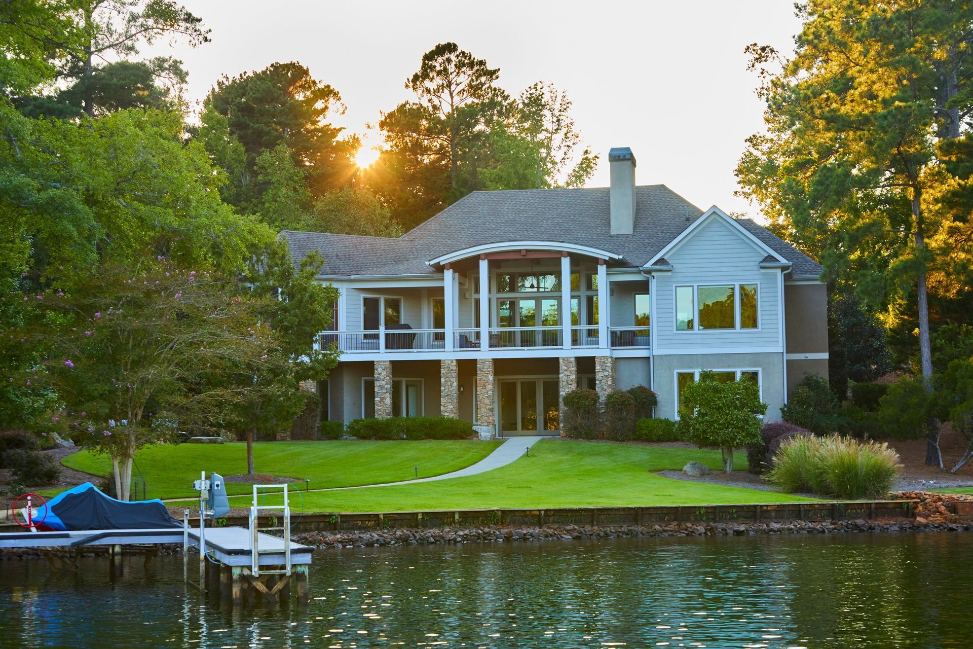 Sunset view of a white lakeside house with a dock and green lawn framed by trees
