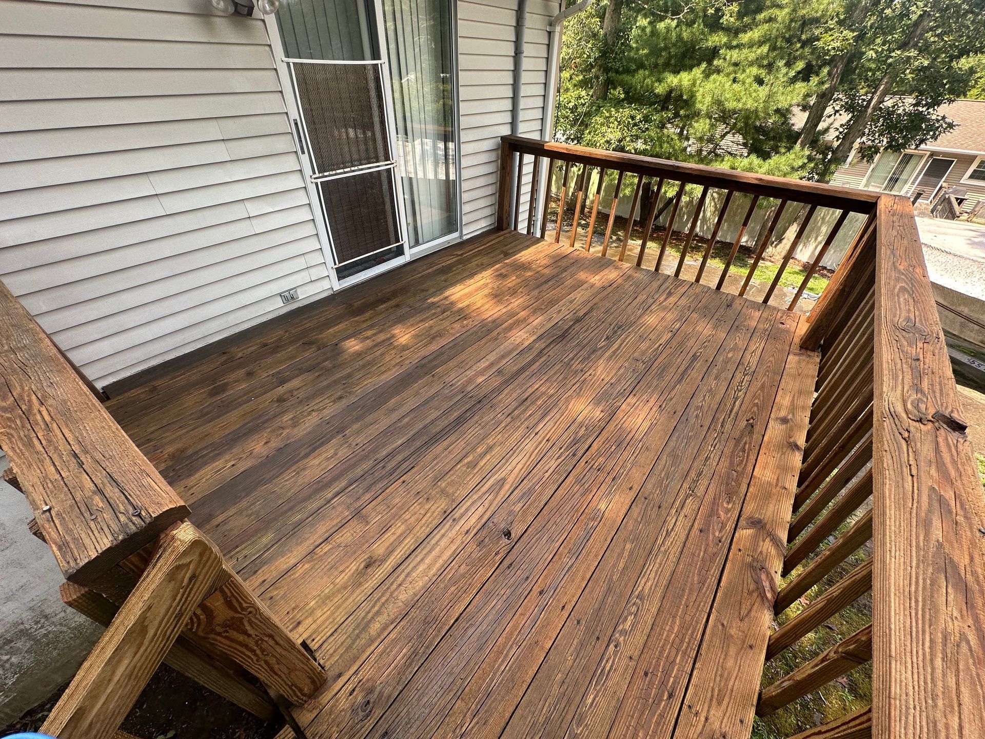 Weathered wooden deck beside a house, with brown railings and sunlight filtering through nearby trees