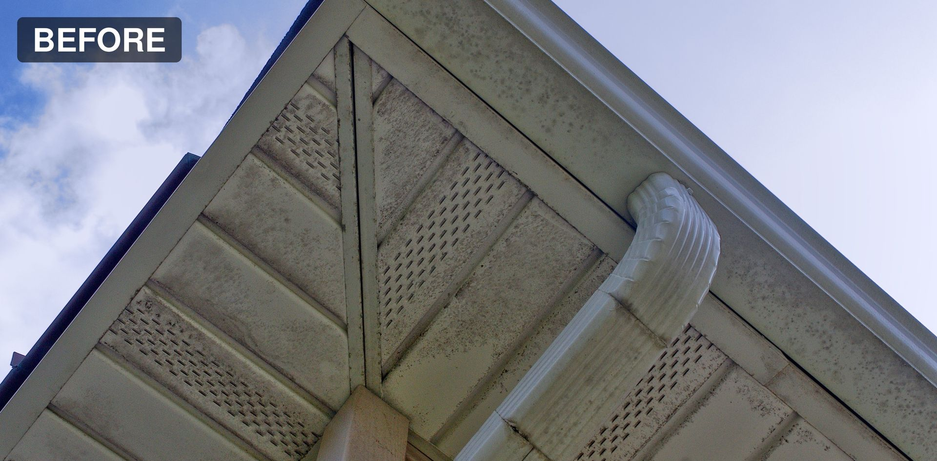 Underside of a roof eave with beige soffit and a white gutter downspout against blue sky