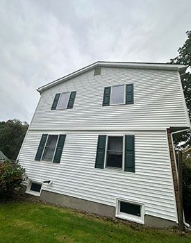White-sided two-story house with dark shutters and basement windows on a grassy slope