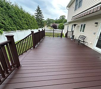 Spacious brown wooden deck beside a white house, with railings, chairs, and a backyard lawn beyond.