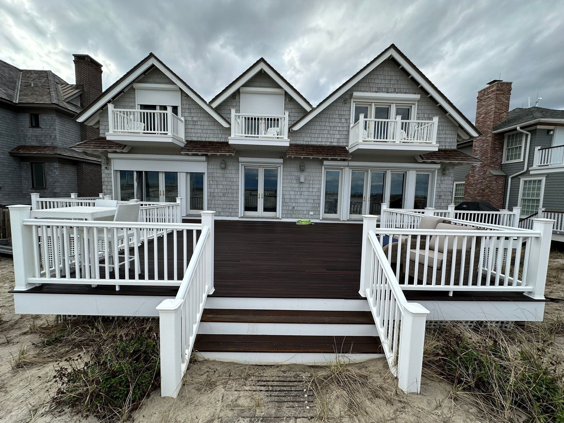Spacious brown wooden deck beside a white house, with railings, chairs, and a backyard lawn beyond.