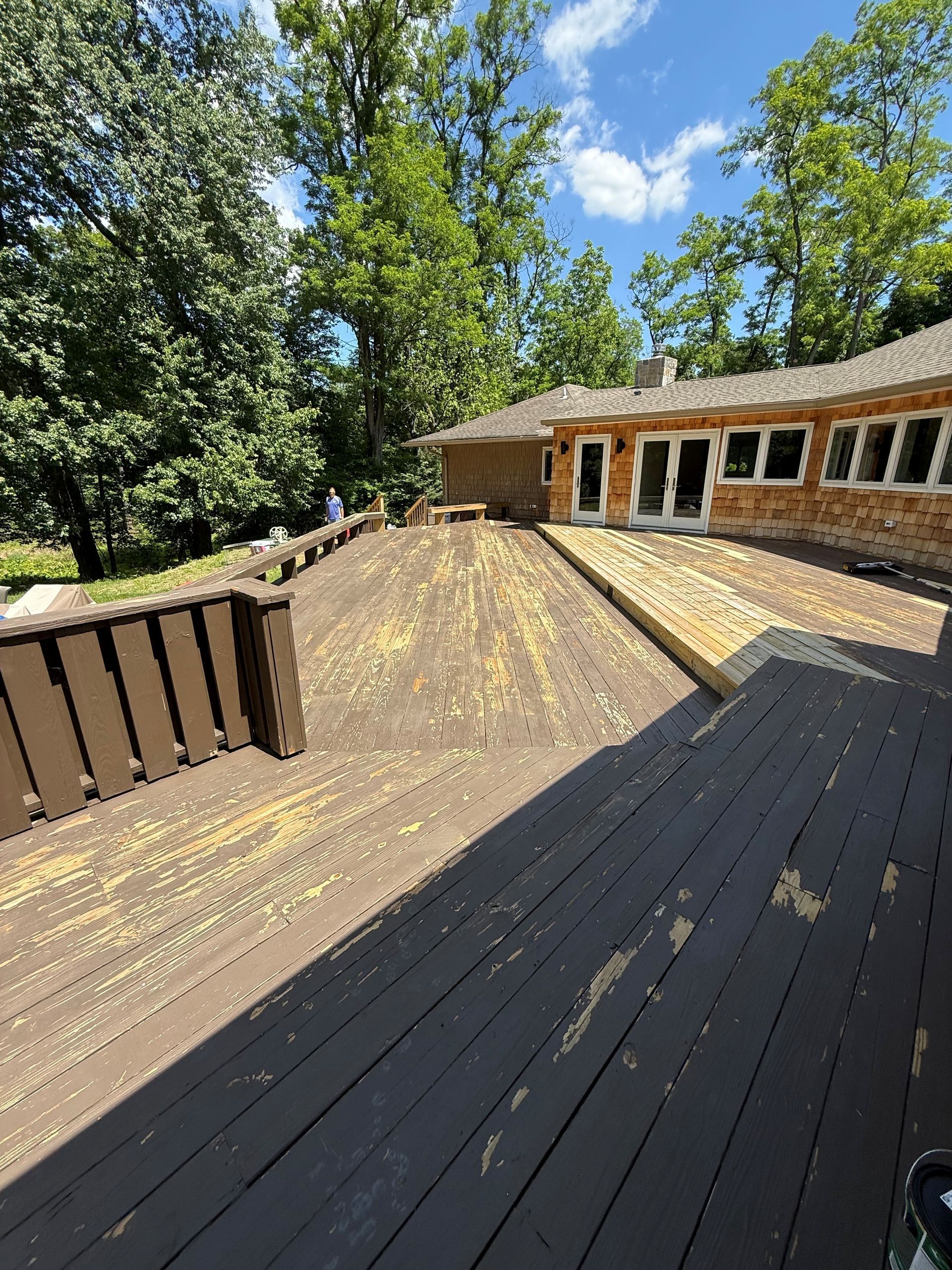 Weathered wooden deck beside a house, with brown railings and sunlight filtering through nearby trees