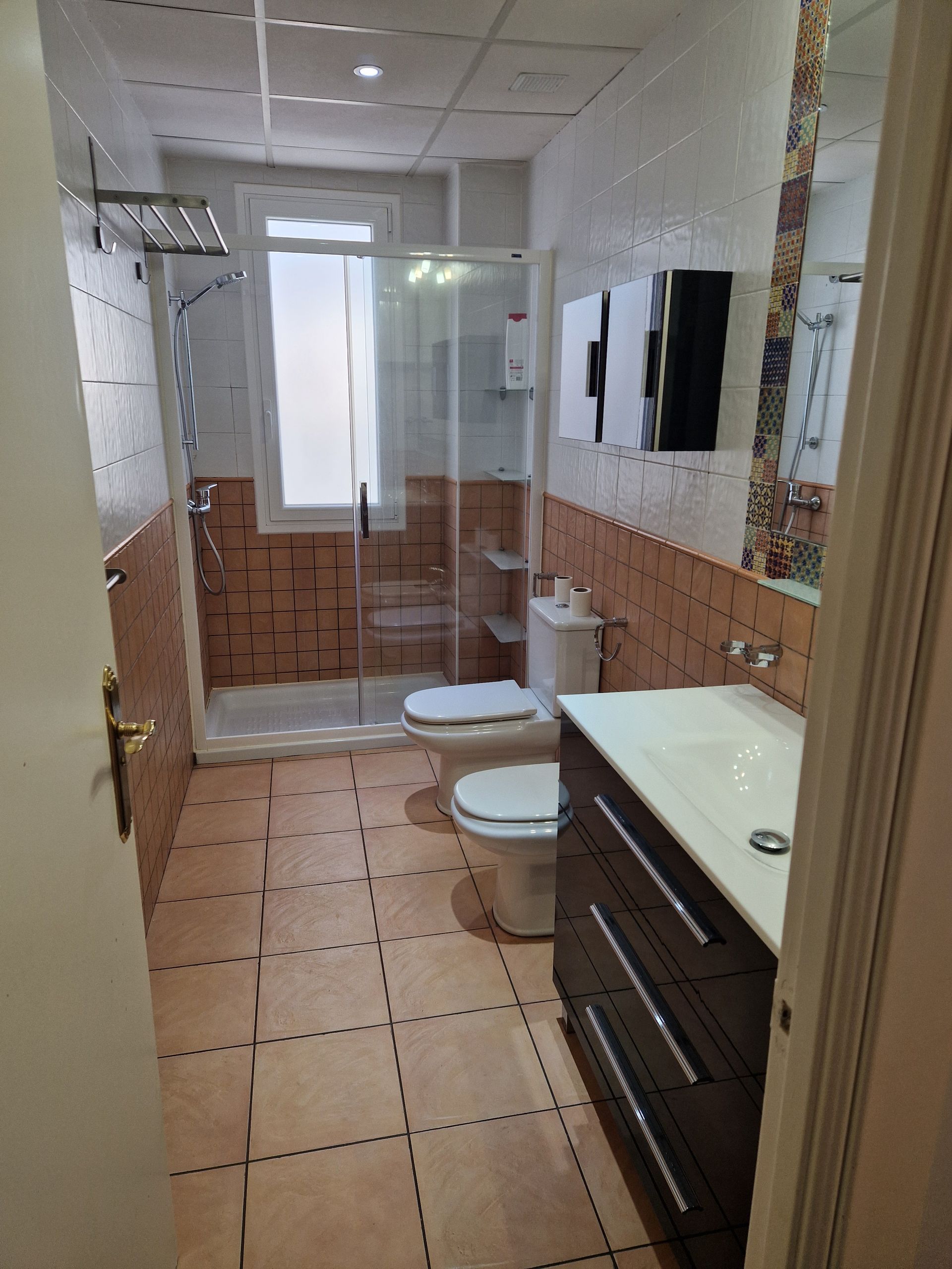 Baño estrecho con azulejos, ducha, inodoro, lavabo con mueble, espejo y una ventana luminosa al final.