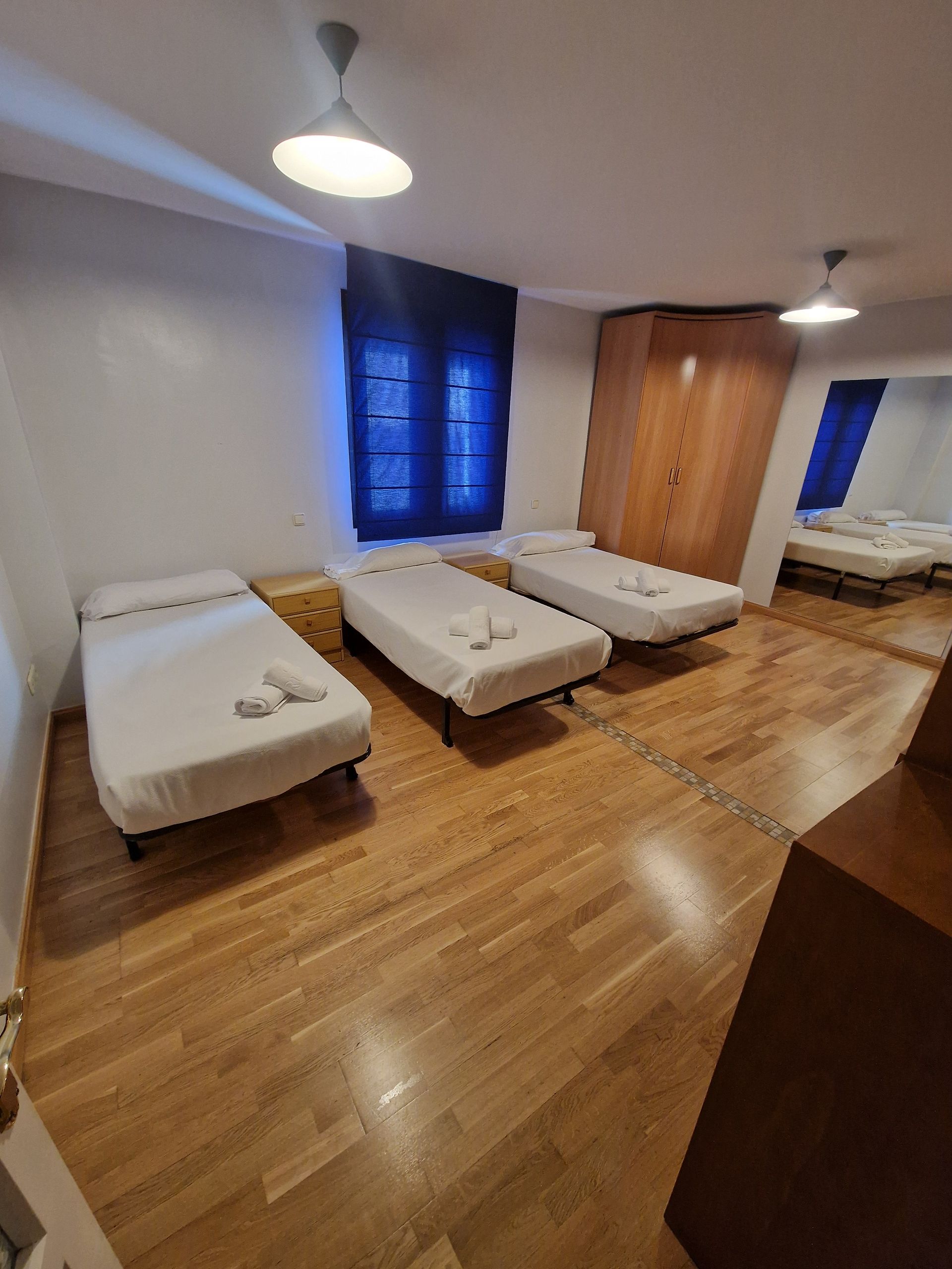 Habitación sencilla con tres camas, suelo de madera, cortinas azules y un armario bajo luces de techo.
