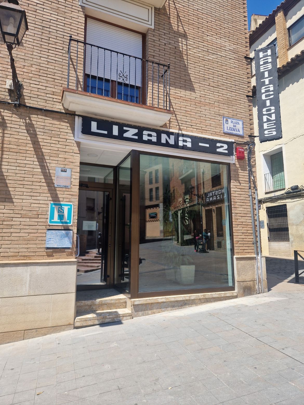 Entrada al local comercial LIZANA-2 en una calle soleada, con puerta de cristal y fachada de ladrillo.