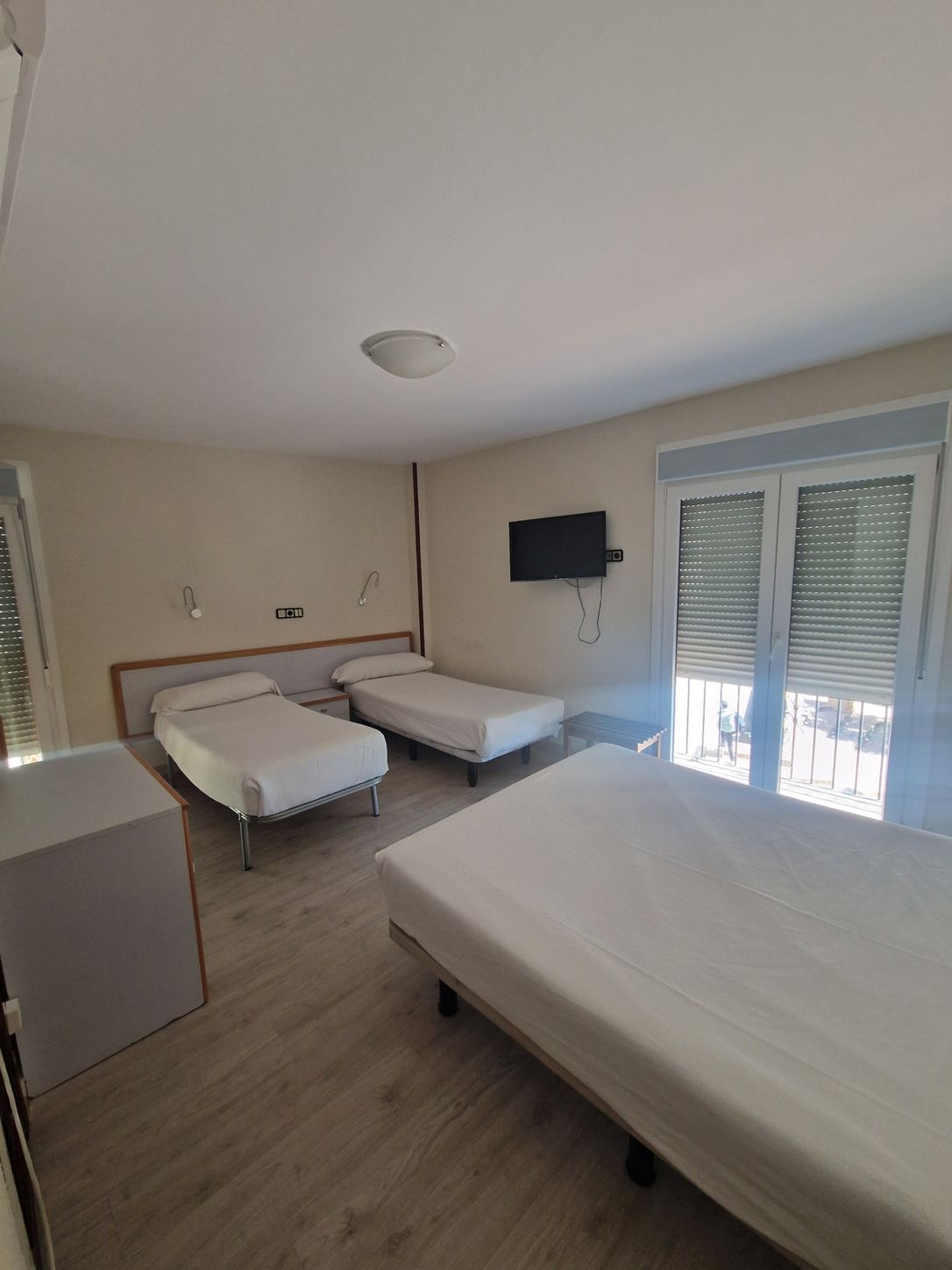 Amplia habitación de hotel con tres camas blancas, televisor en la pared y puertas de balcón con contraventanas.