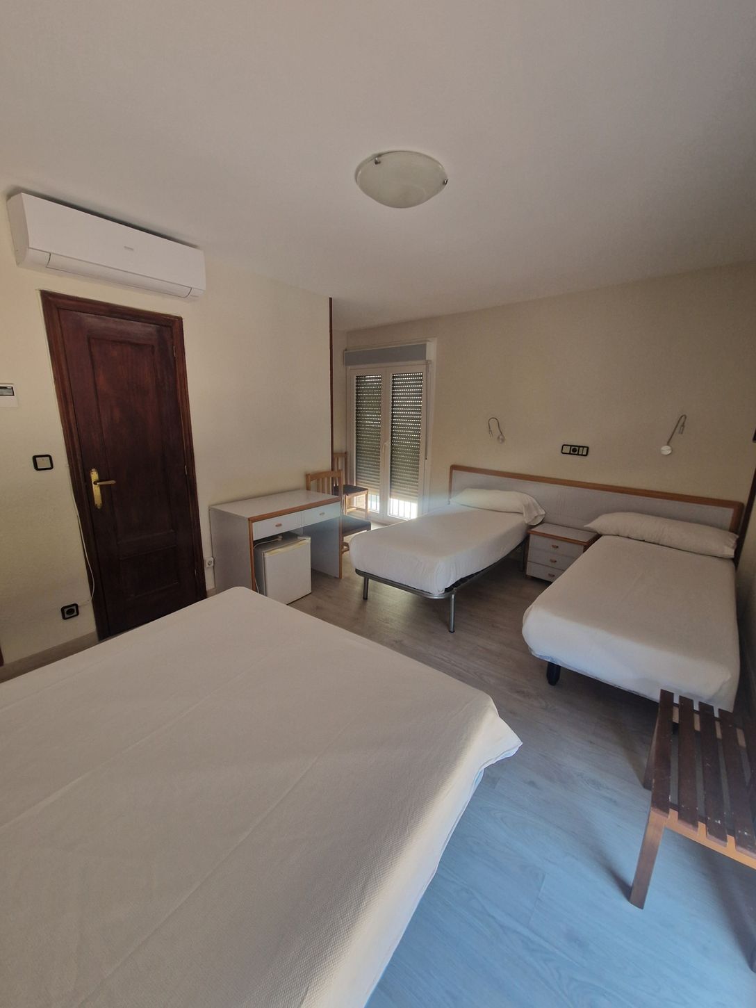 Habitación de hotel pequeña con tres camas individuales, puerta de madera, escritorio y contraventanas.