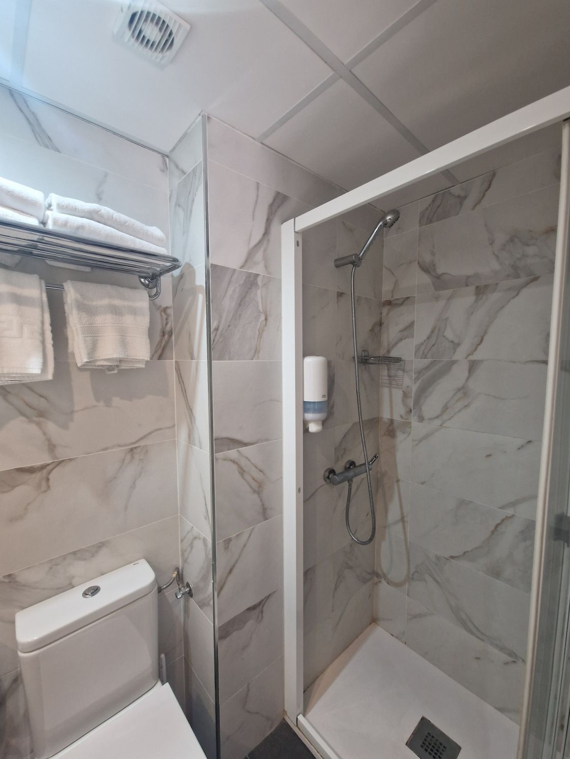 Baño pequeño con ducha, azulejos que imitan el mármol, mampara de cristal, inodoro y ventilador de techo.