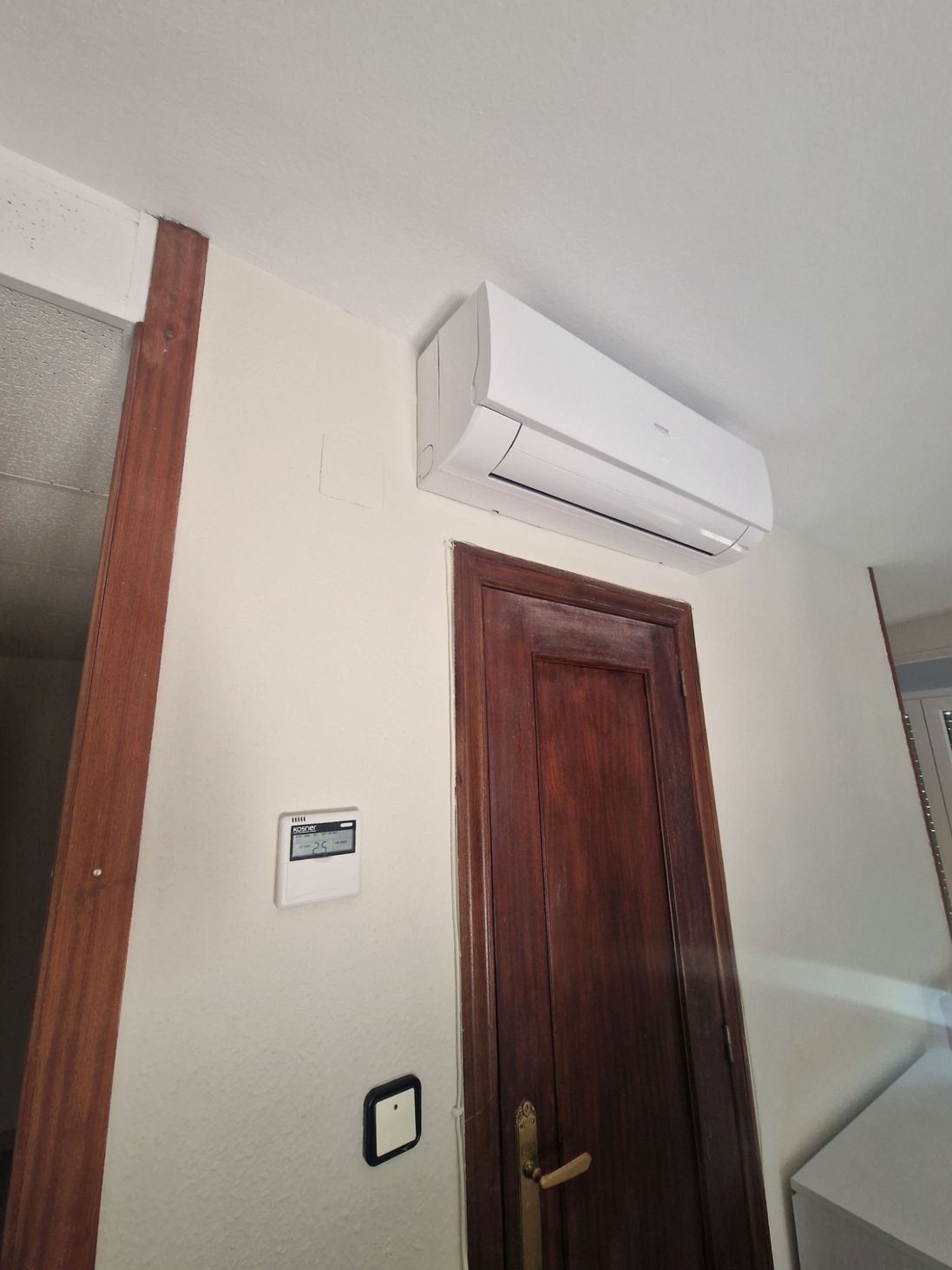 Pasillo con un aire acondicionado blanco montado en la pared, encima de una puerta de madera y un interruptor de luz.