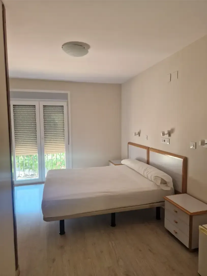 Ventana abierta en el dormitorio con balcón y vistas a edificios y árboles de colores pastel; cama blanca en primer plano.