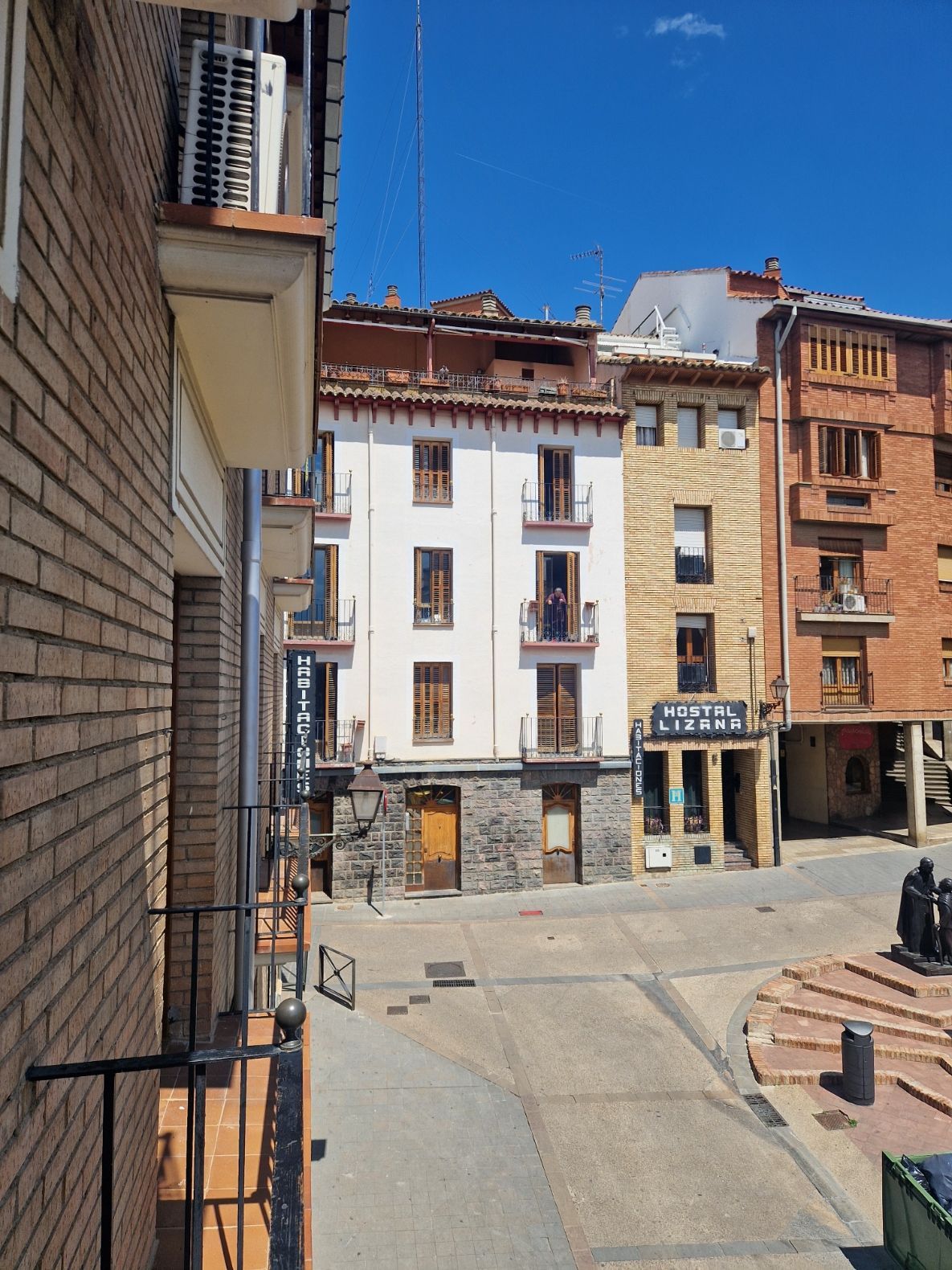 Plaza europea soleada con edificios de ladrillo y blancos, una calle estrecha y un cielo azul despejado.
