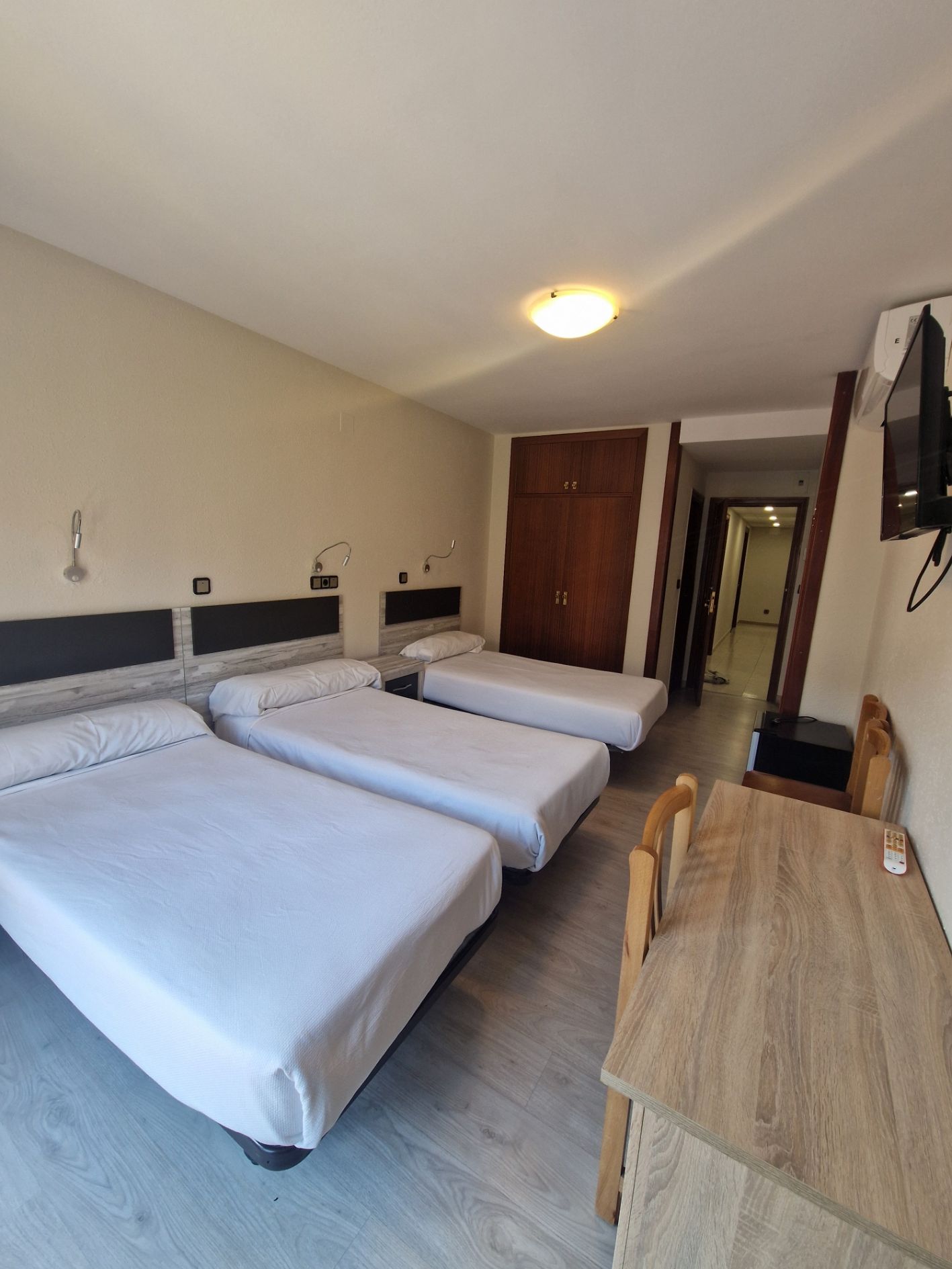 Habitación de hotel con tres camas individuales, escritorio de madera, televisor montado en la pared y una ventana junto a la puerta.