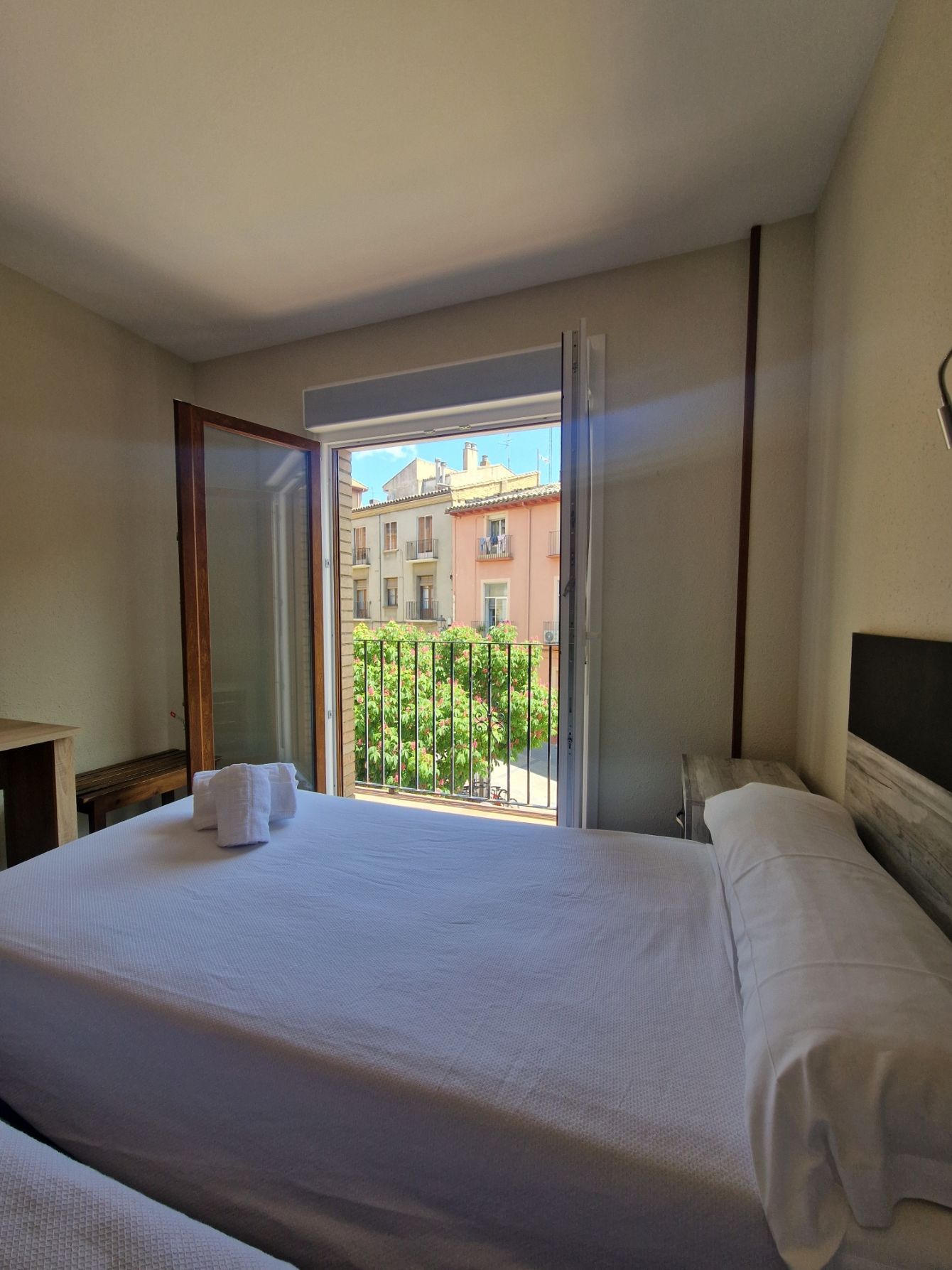 Habitación pequeña con una cama junto a una ventana abierta con vistas a una calle y edificios.