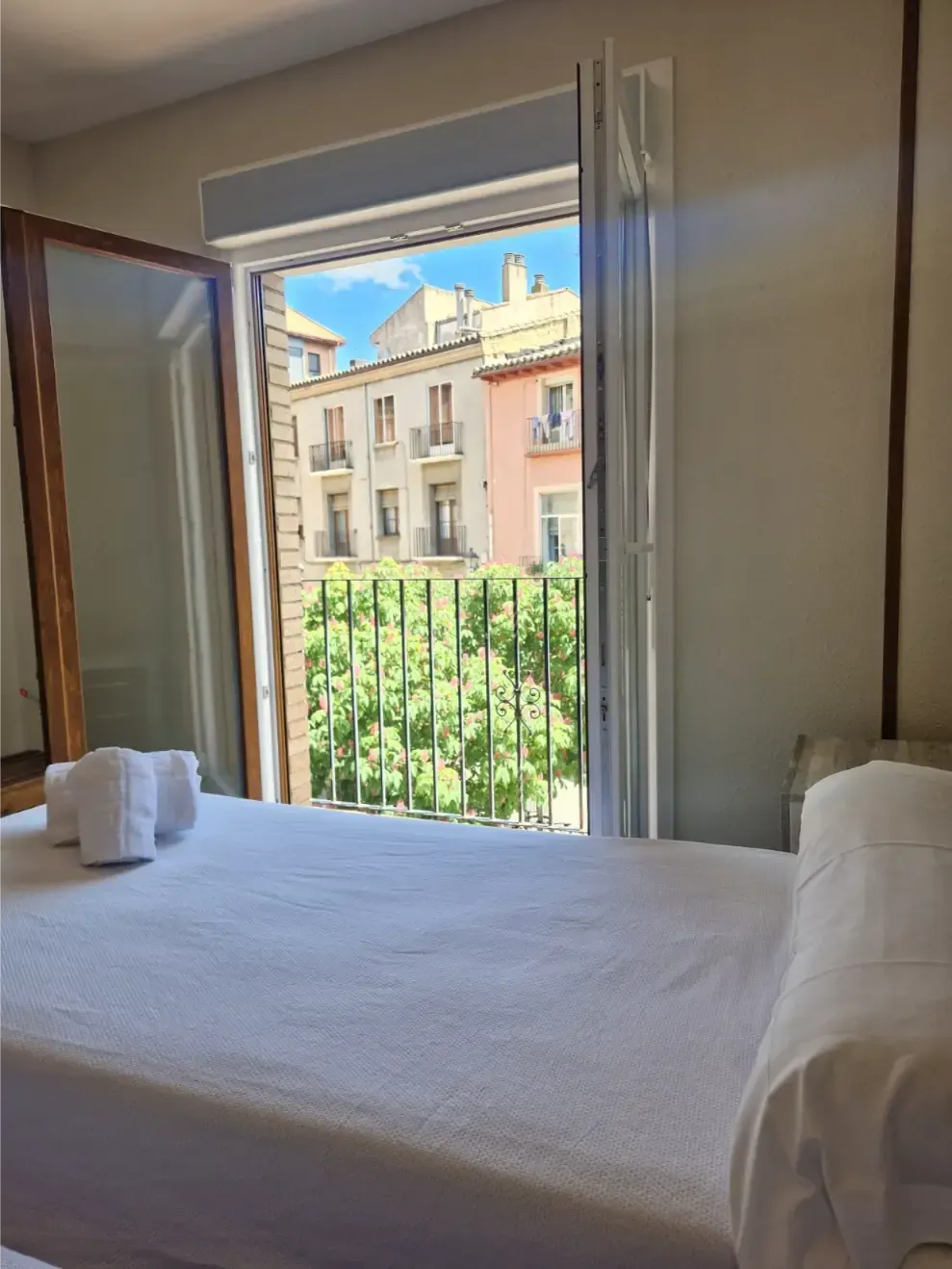 Ventana abierta en el dormitorio con balcón y vistas a edificios y árboles de colores pastel; cama blanca en primer plano.