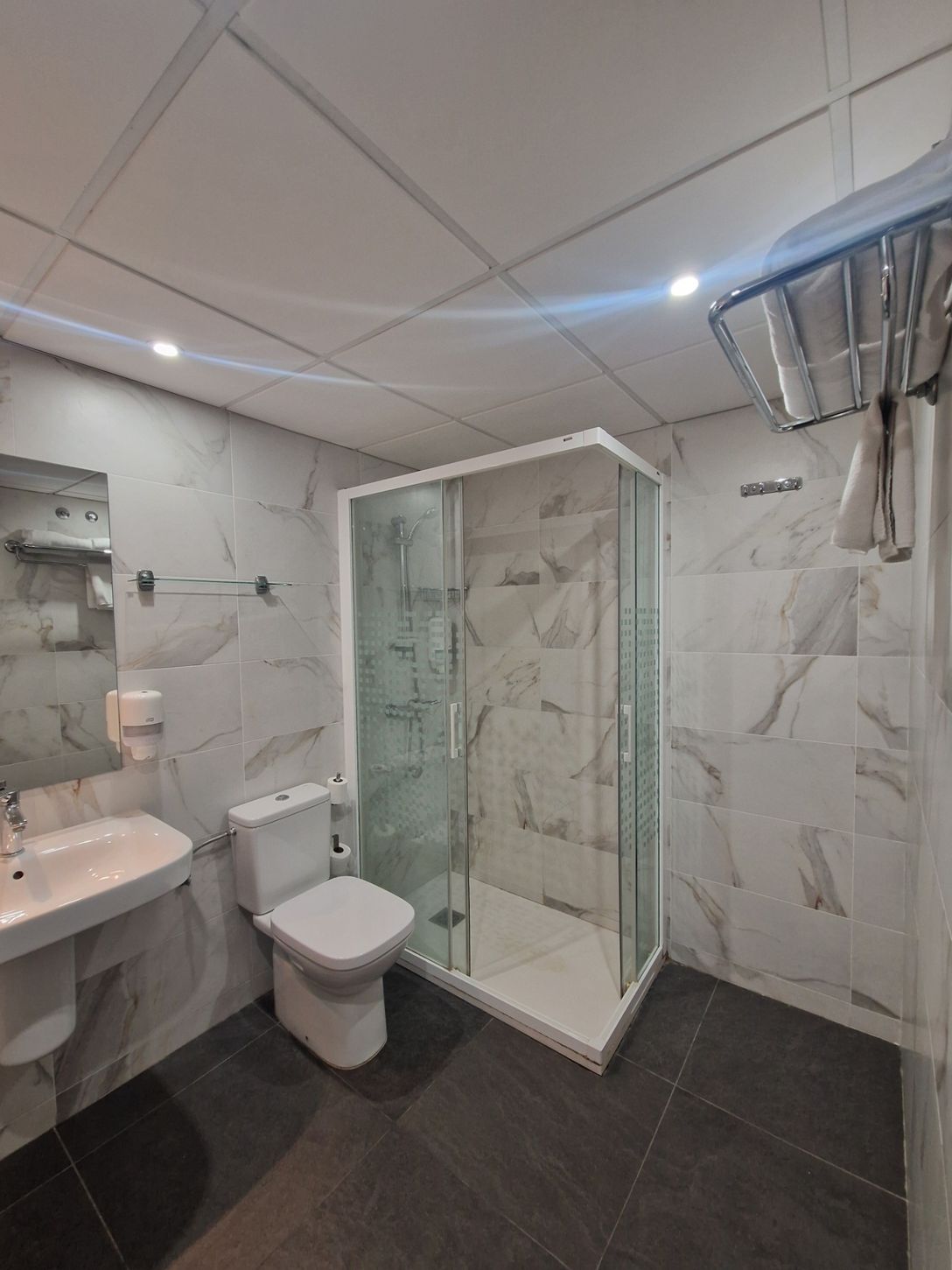 Baño moderno con ducha de cristal, inodoro, lavabo, paredes de mármol y suelo de baldosas oscuras.