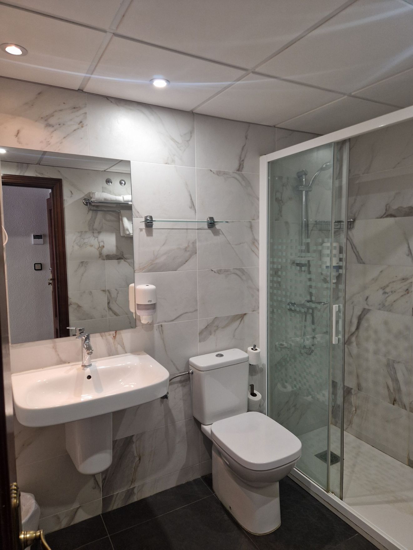 Baño moderno con lavabo, inodoro, ducha de cristal y espejo de pared; azulejos blancos estilo mármol e iluminación empotrada.