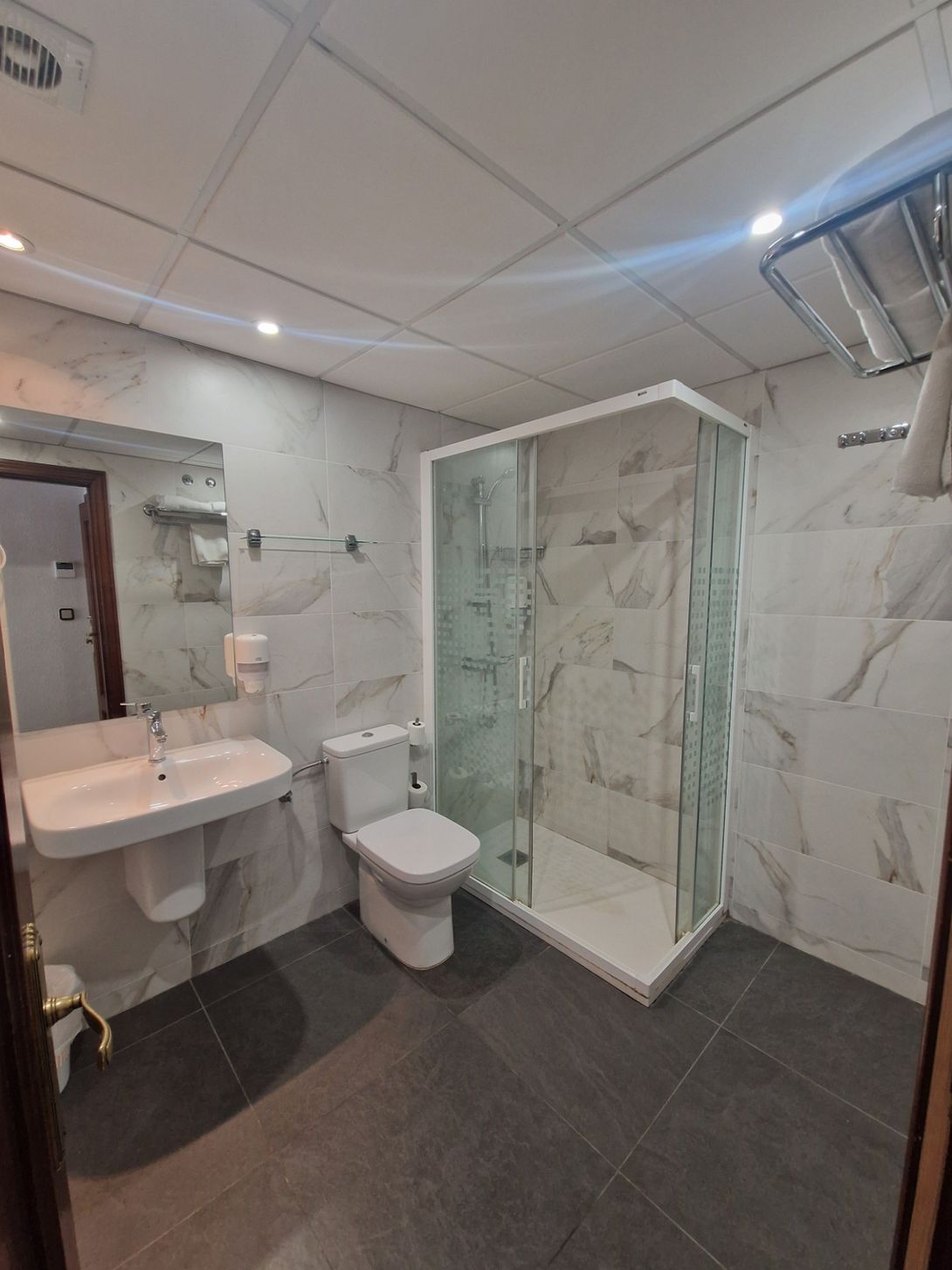 Baño moderno con paredes de mármol blanco, cabina de ducha de cristal, lavabo suspendido, inodoro y suelo de baldosas oscuras.