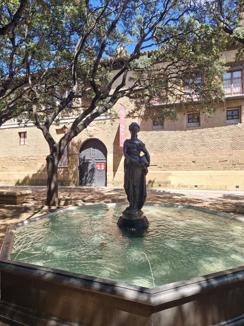 Fuente de piedra con una estatua en un patio soleado junto a un edificio histórico, a la sombra de un árbol.