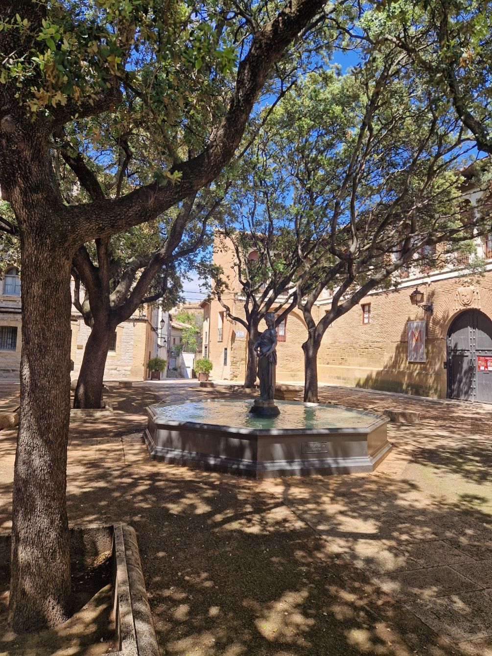 Patio de piedra sombreado con una fuente central, árboles y muros de ladrillo históricos bajo un cielo azul.