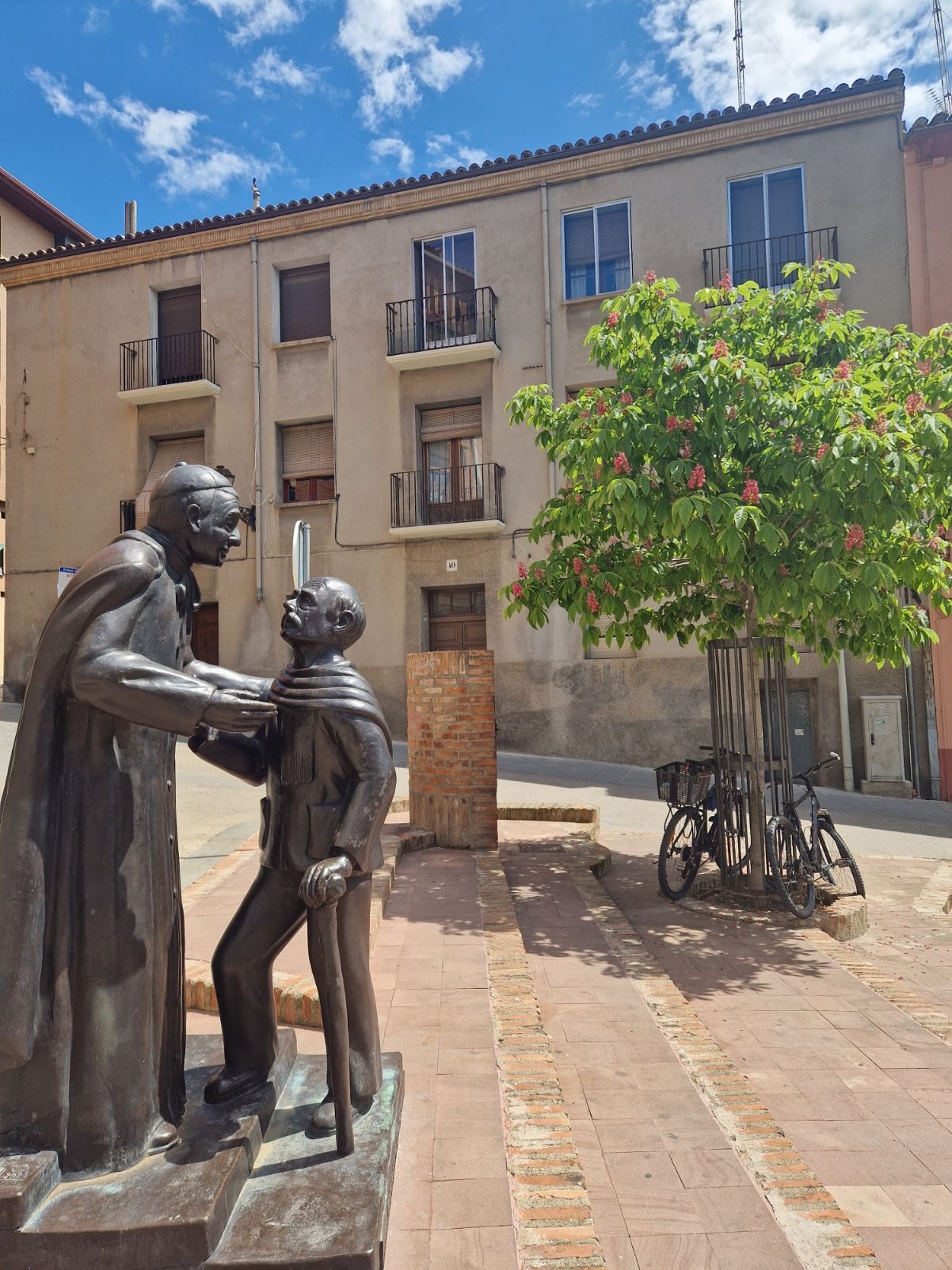 Estatua de bronce de dos personas en una plaza soleada, con un árbol y edificios de apartamentos de color beige al fondo.